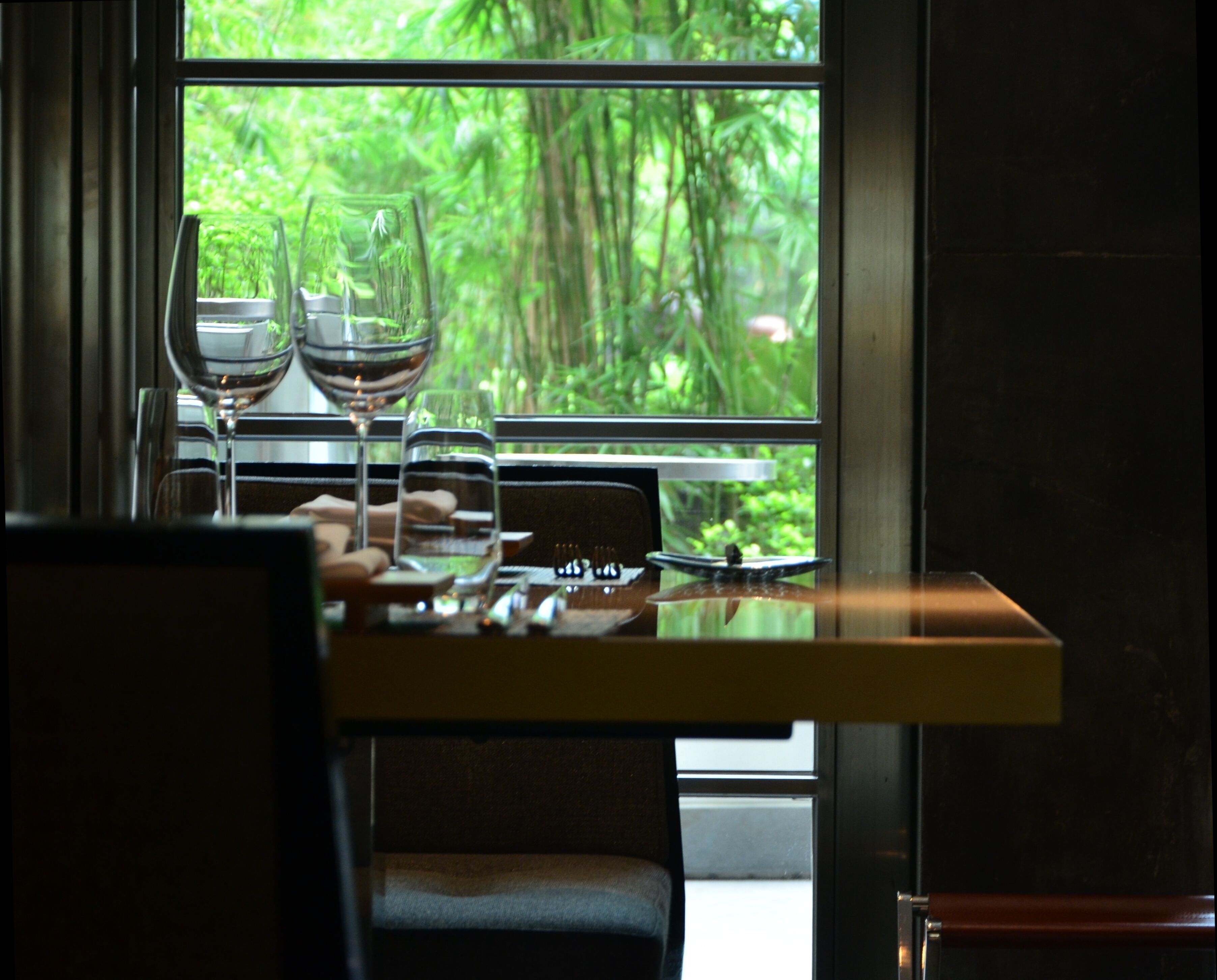A table with two wine glasses and a water glass, set against a window with a view of dense greenery.