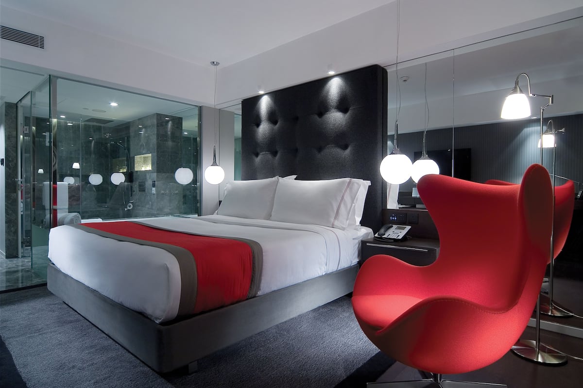 A modern hotel room with a large bed, a red chair, and a glass shower enclosure.