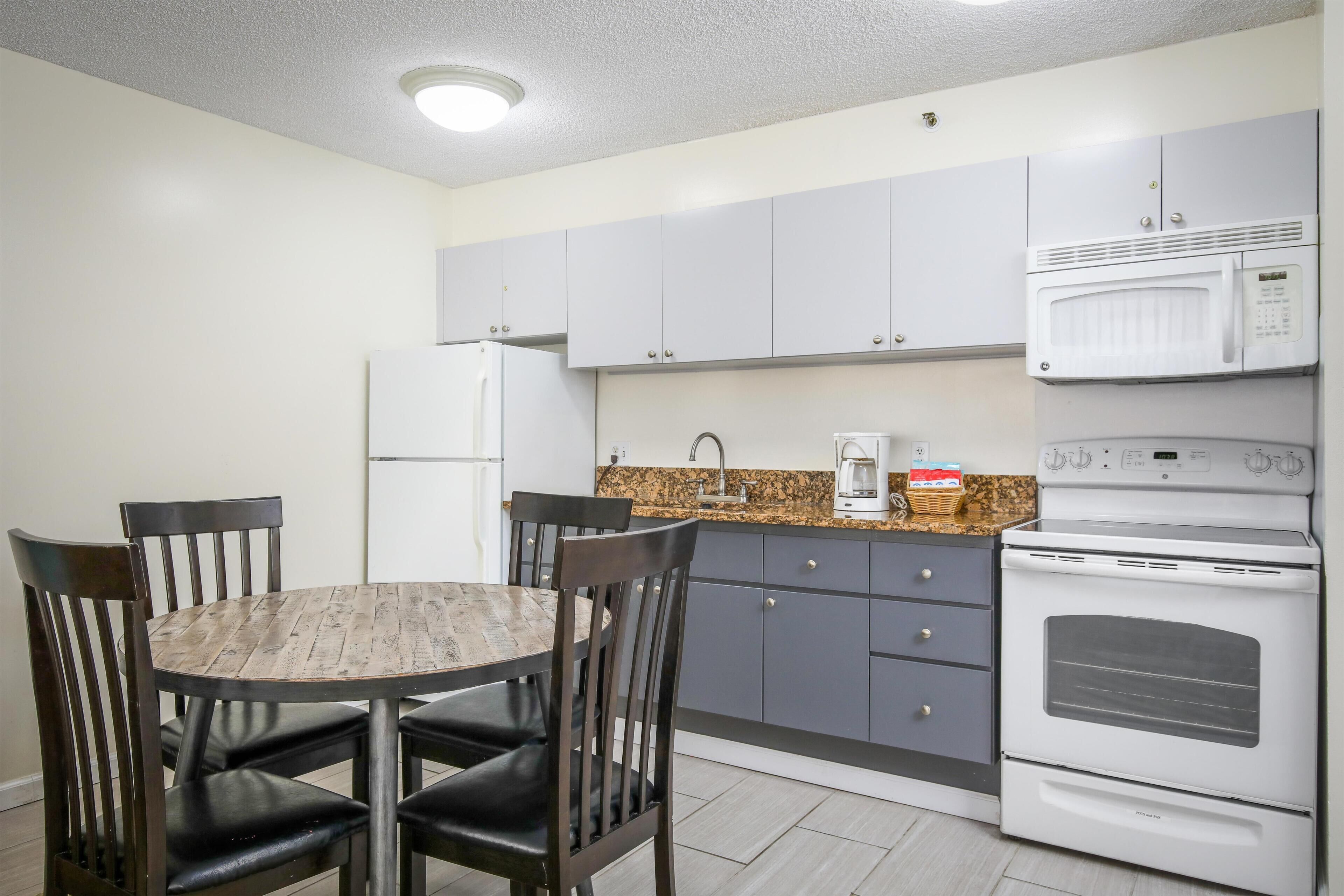 A compact kitchen with a round wooden table, four chairs, a refrigerator, a microwave, a stove, and a sink.