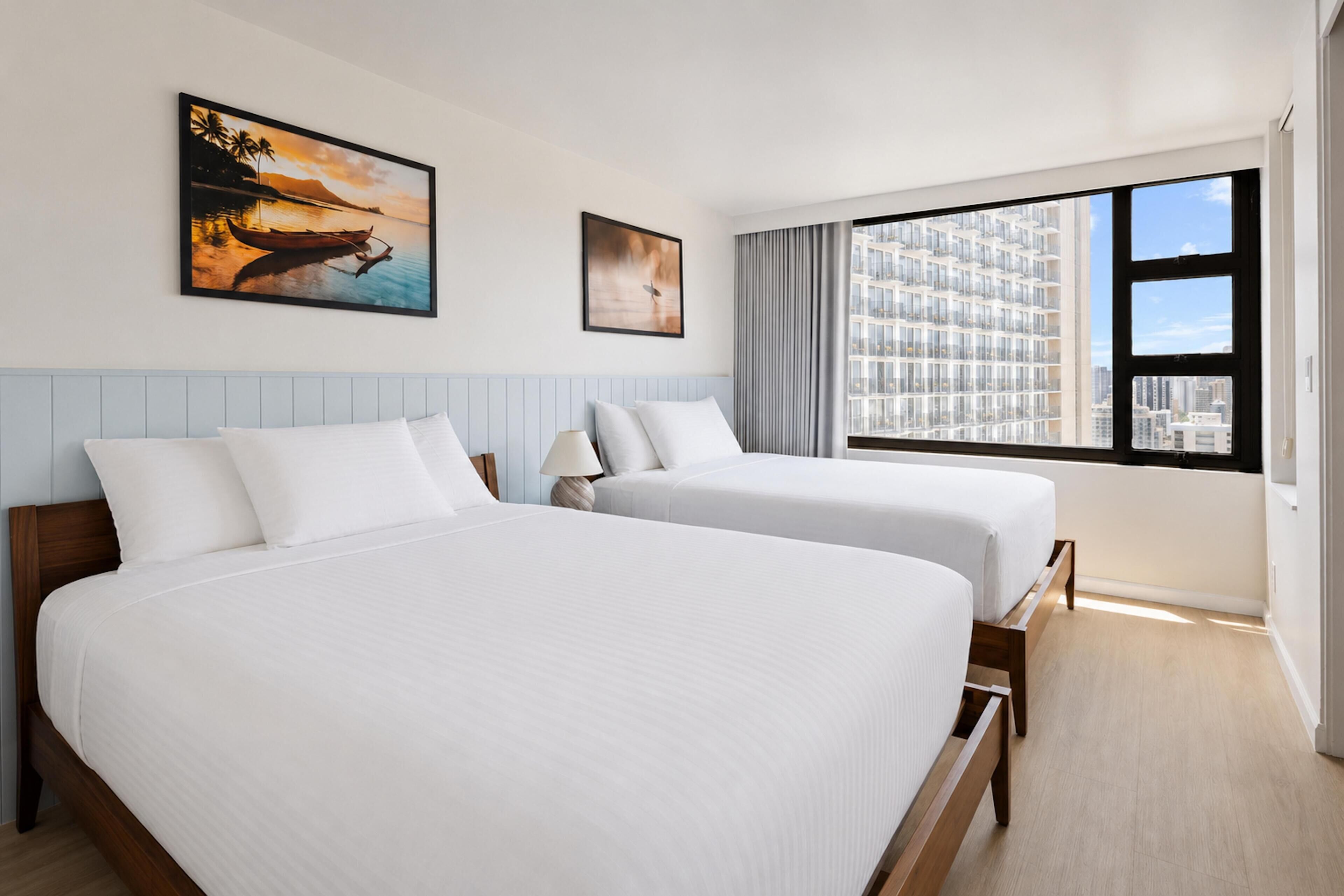 A hotel room with two beds, a large window with a city view, and two framed pictures on the wall.