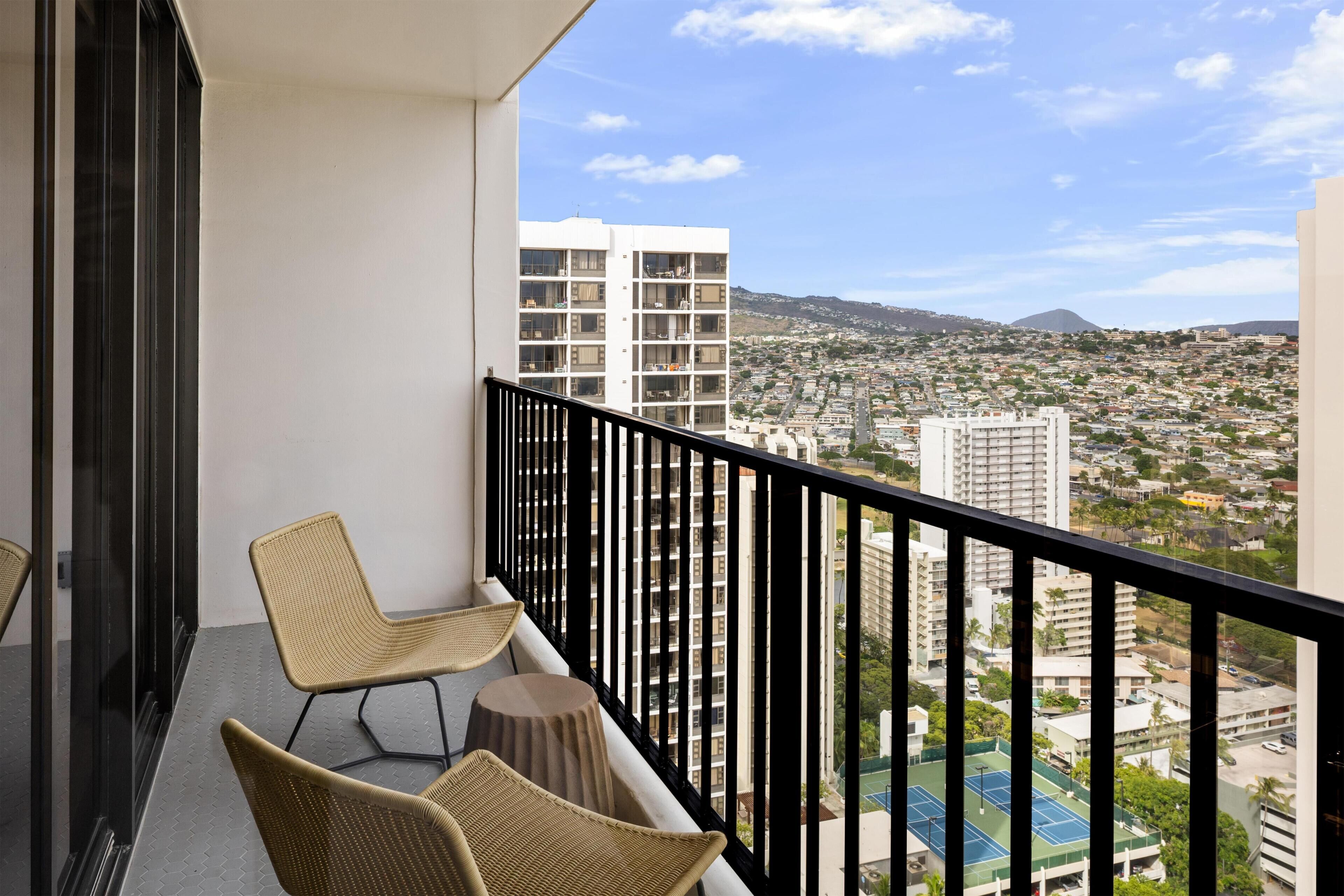 A balcony with two wicker chairs and a small round table, overlooking a densely packed urban area with buildings and a distant mountain.