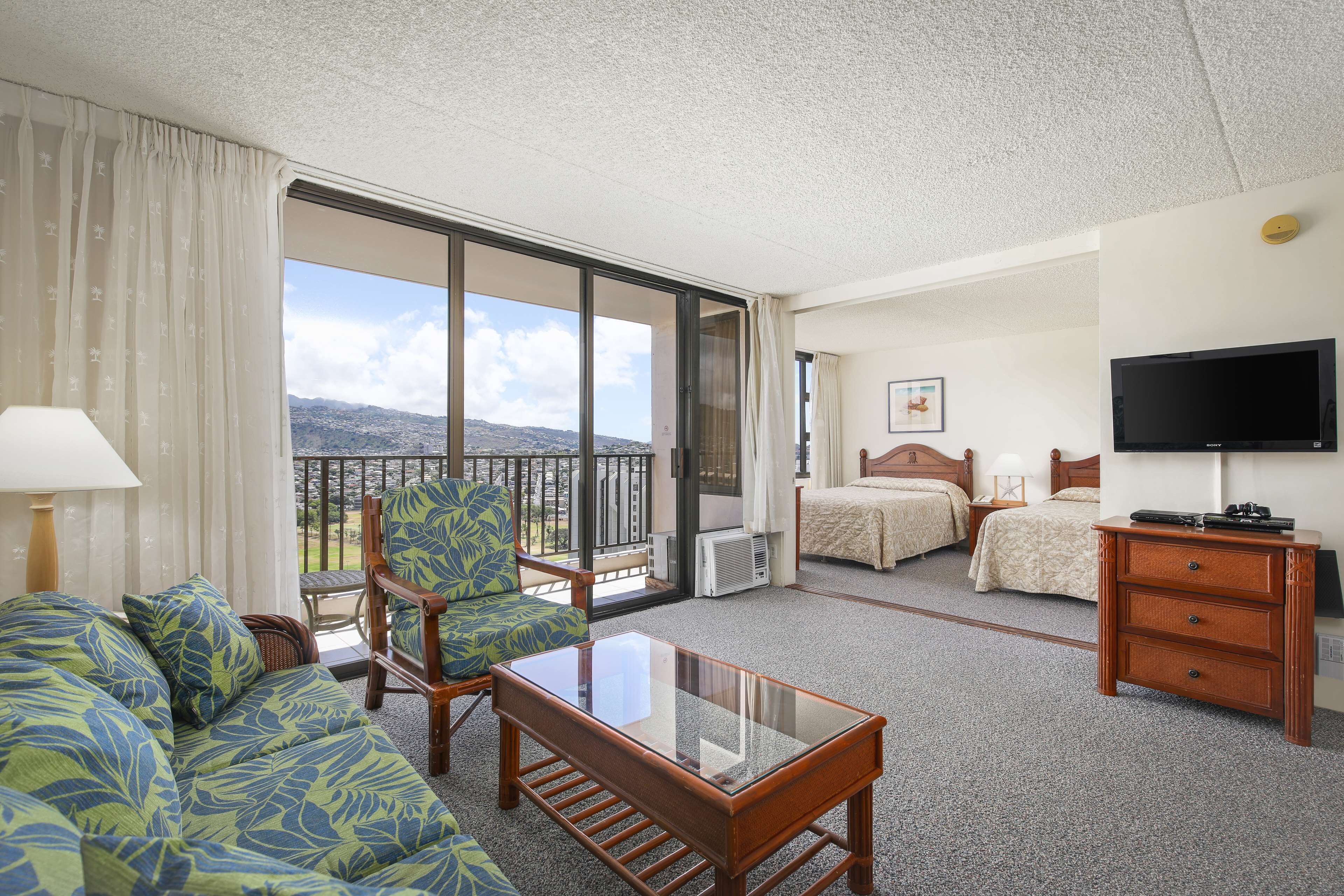 A hotel room with a balcony, a sofa, a chair, a bed, a TV, and a dresser.
