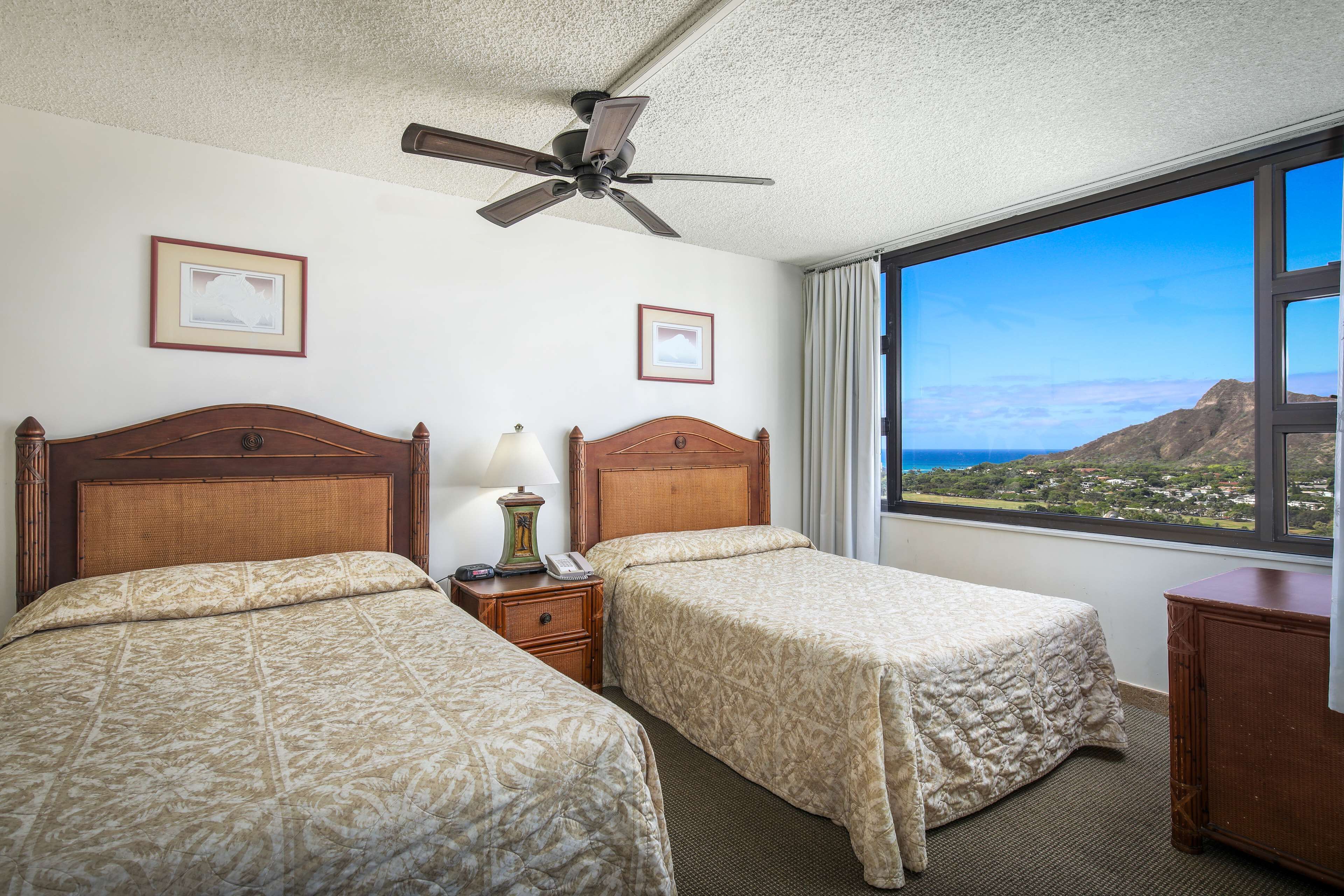 A hotel room with two beds, a ceiling fan, a nightstand with a lamp, and a large window offering a scenic view.