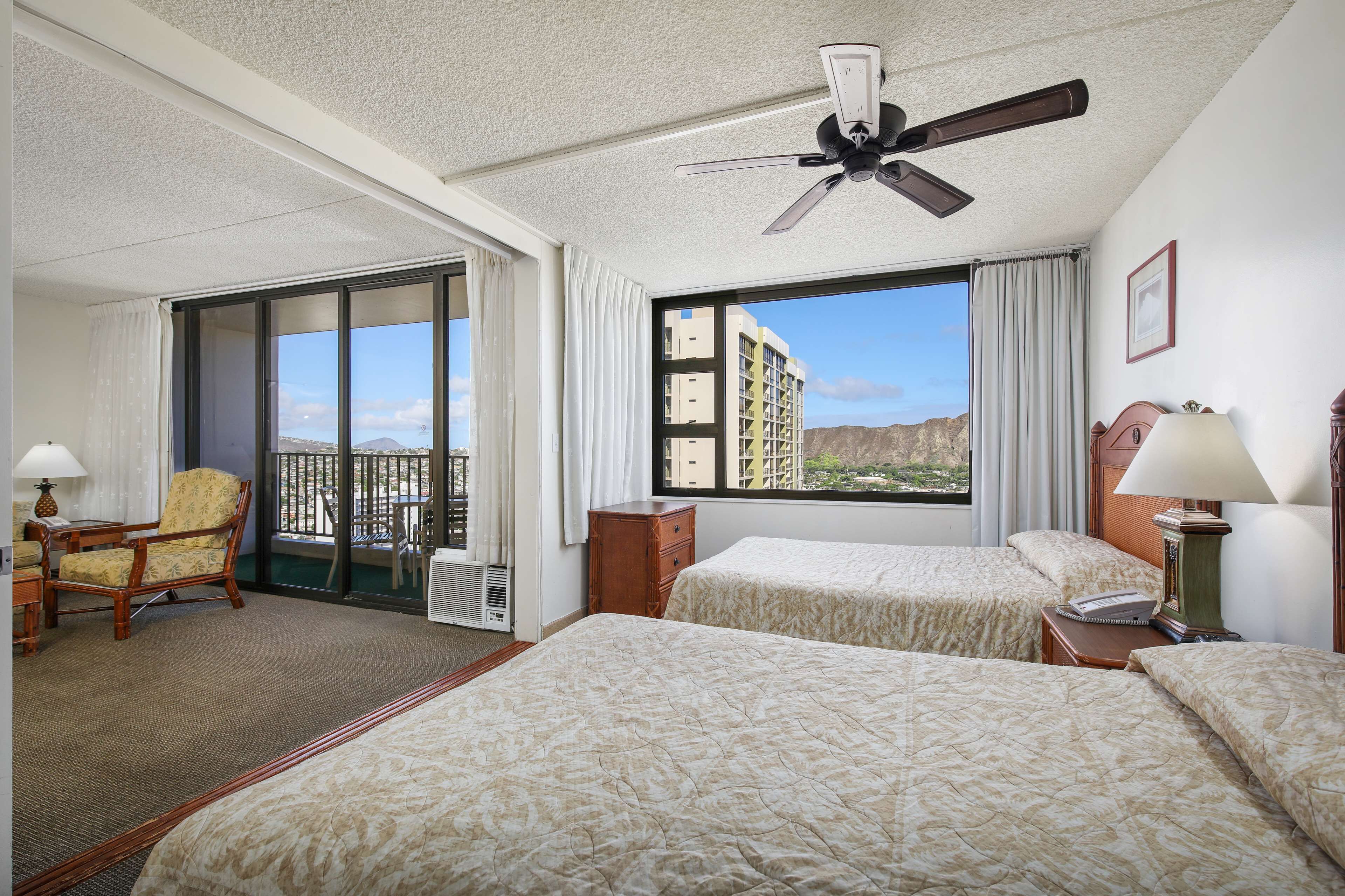 A hotel room with two beds, a chair, a desk, a lamp, a ceiling fan, and a large window with a view of buildings and mountains.