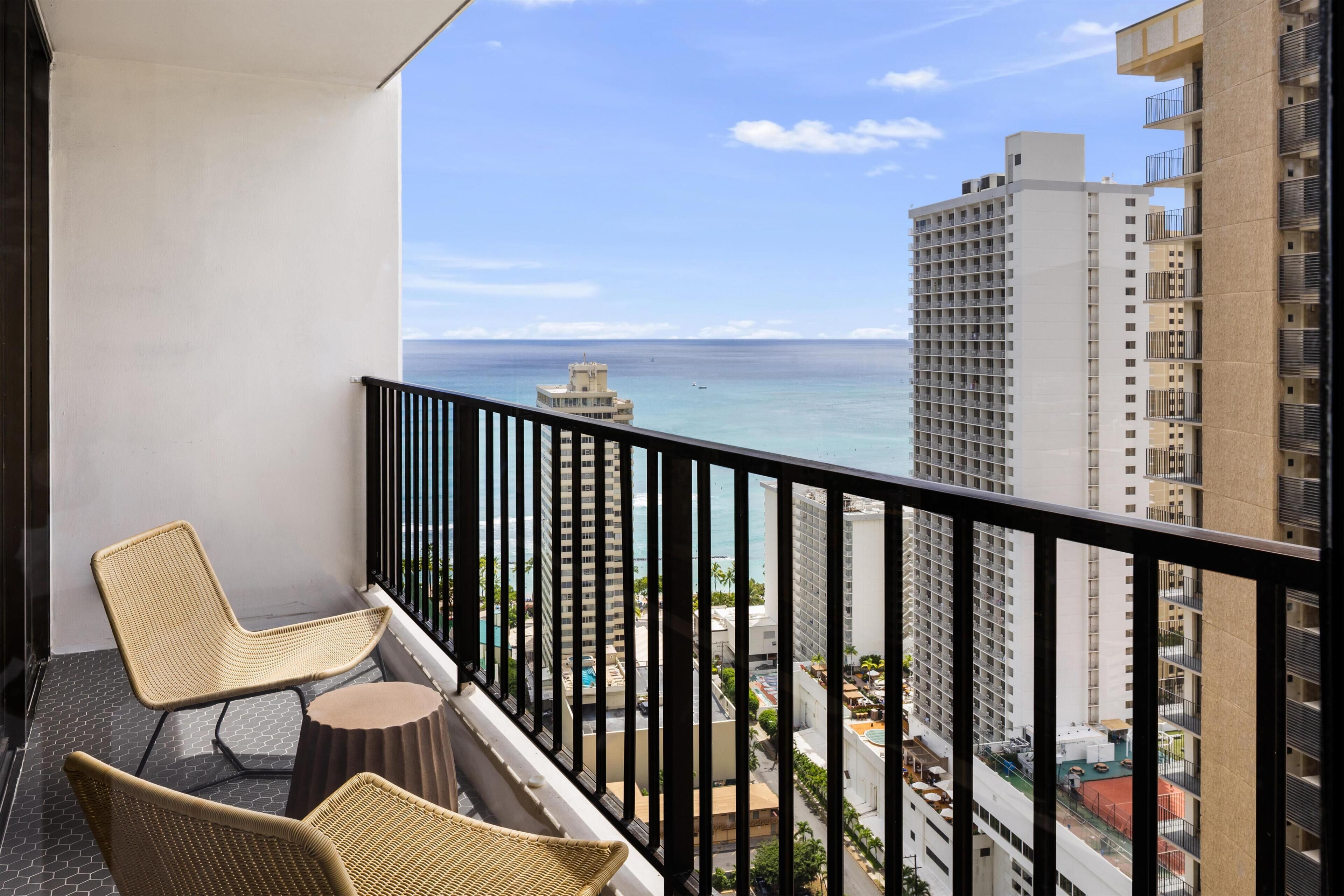 A balcony with two wicker chairs and a small round table, overlooking a cityscape with tall buildings and a view of the ocean.