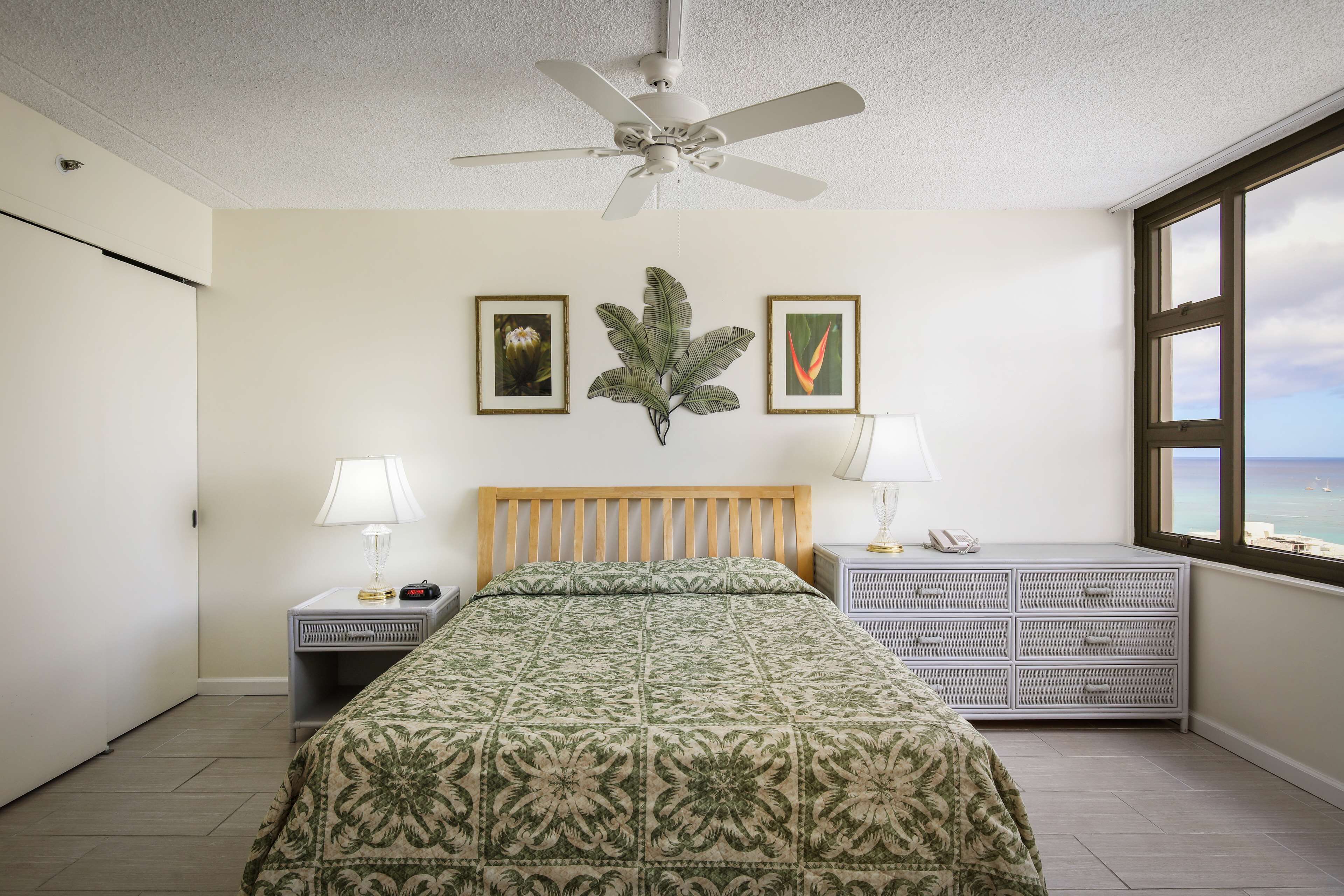A bedroom with a bed, a nightstand, a dresser, a ceiling fan, and a window with a view.
