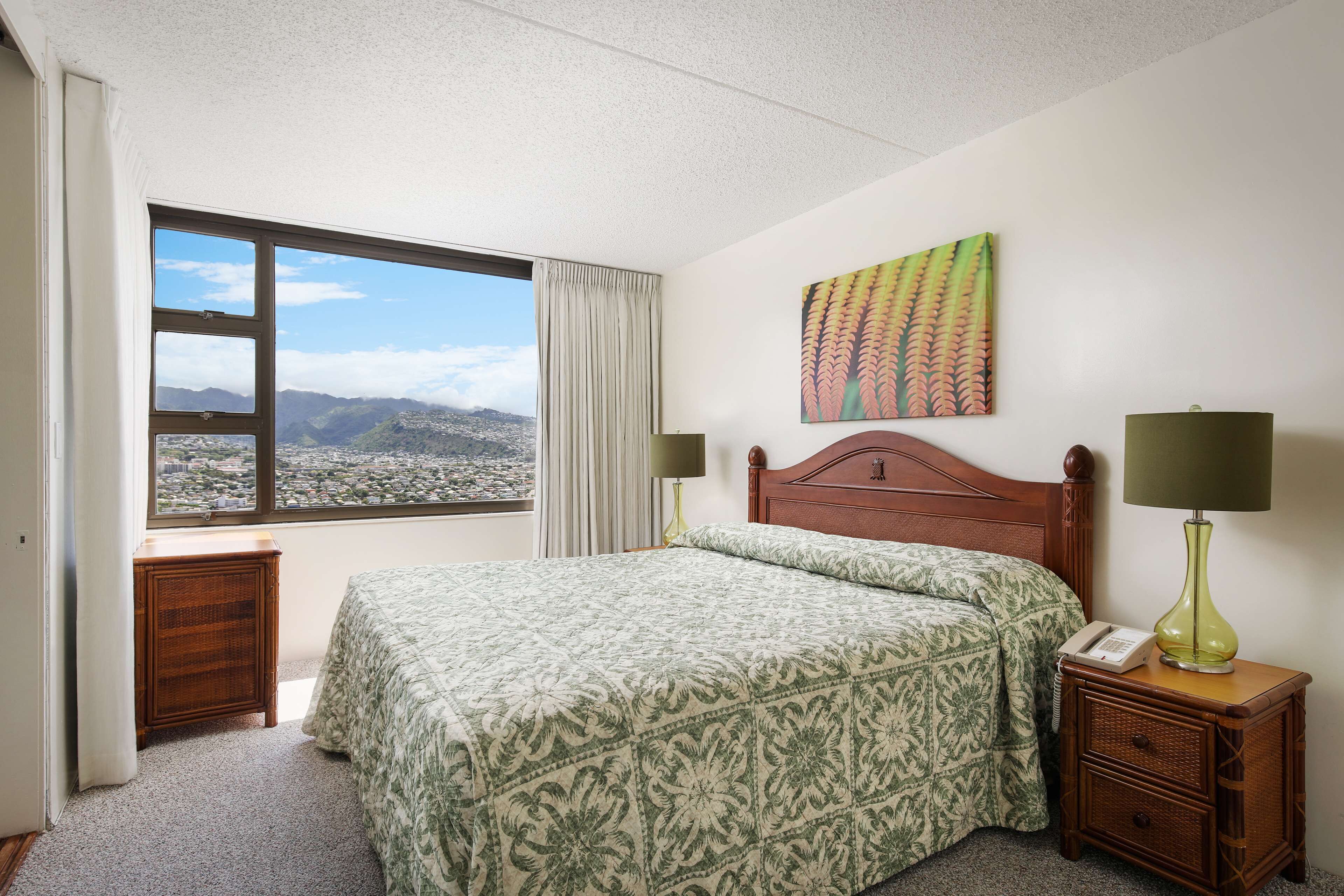 Habitación de hotel con cama, mesita de noche, lámpara, teléfono, ventana con vistas a montañas y un cuadro en la pared.