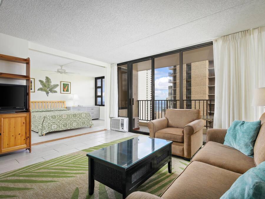 Una sala de estar con sofá, mesa de centro, televisor, cama, balcón con vista a edificios y ventilador de techo.