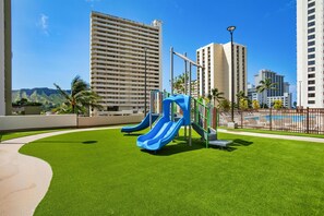 A playground with slides, surrounded by a well-maintained lawn and tall buildings in the background. - Aston at the Waikiki Banyan (Honolulu)
