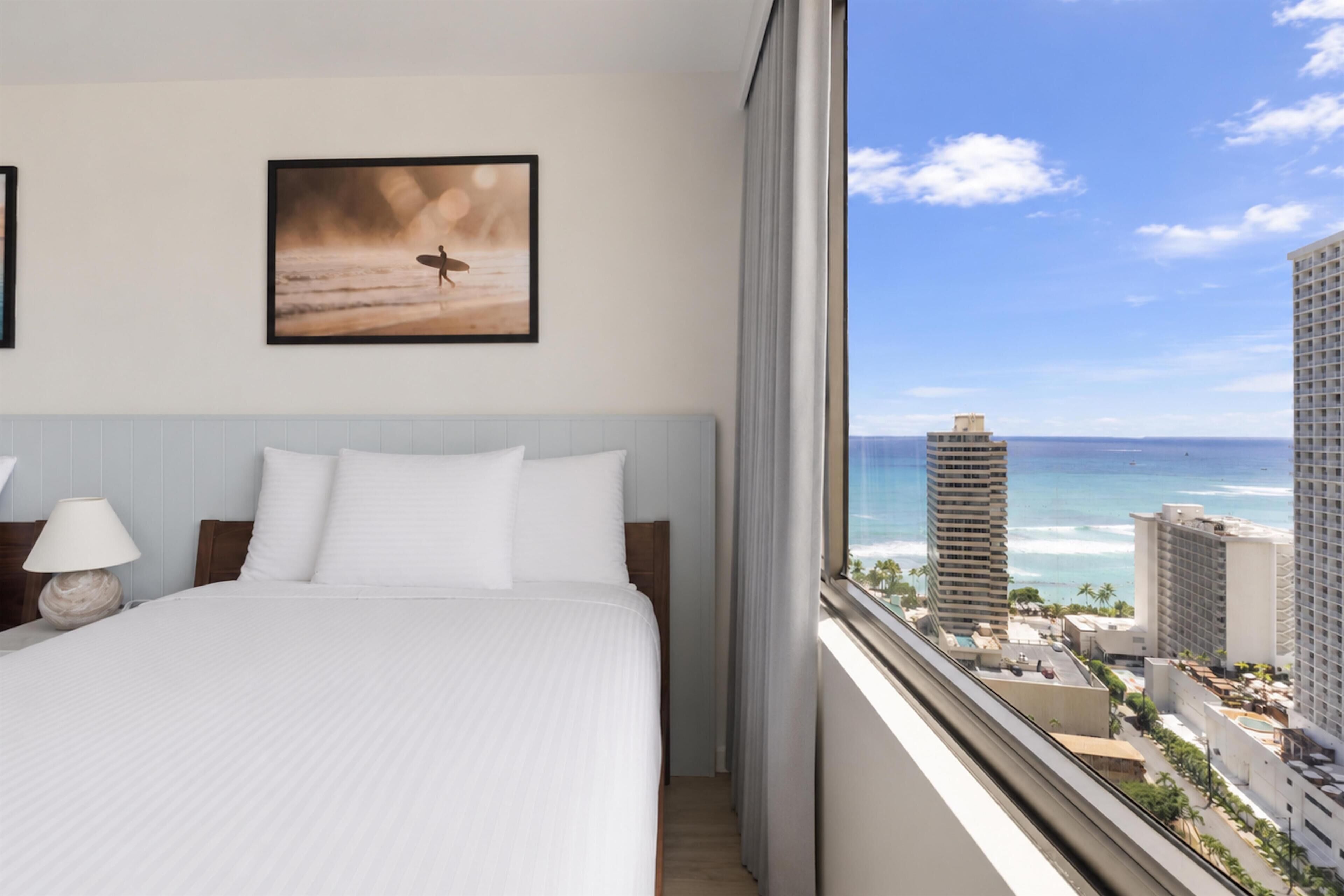 A hotel room with a bed, a framed picture on the wall, and a view of the ocean and buildings.