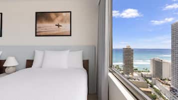 A hotel room with a bed, a framed picture on the wall, and a view of the ocean and buildings.