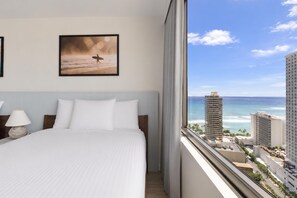 A hotel room with a bed, a framed picture on the wall, and a view of the ocean and buildings. - Aston at the Waikiki Banyan (Honolulu)