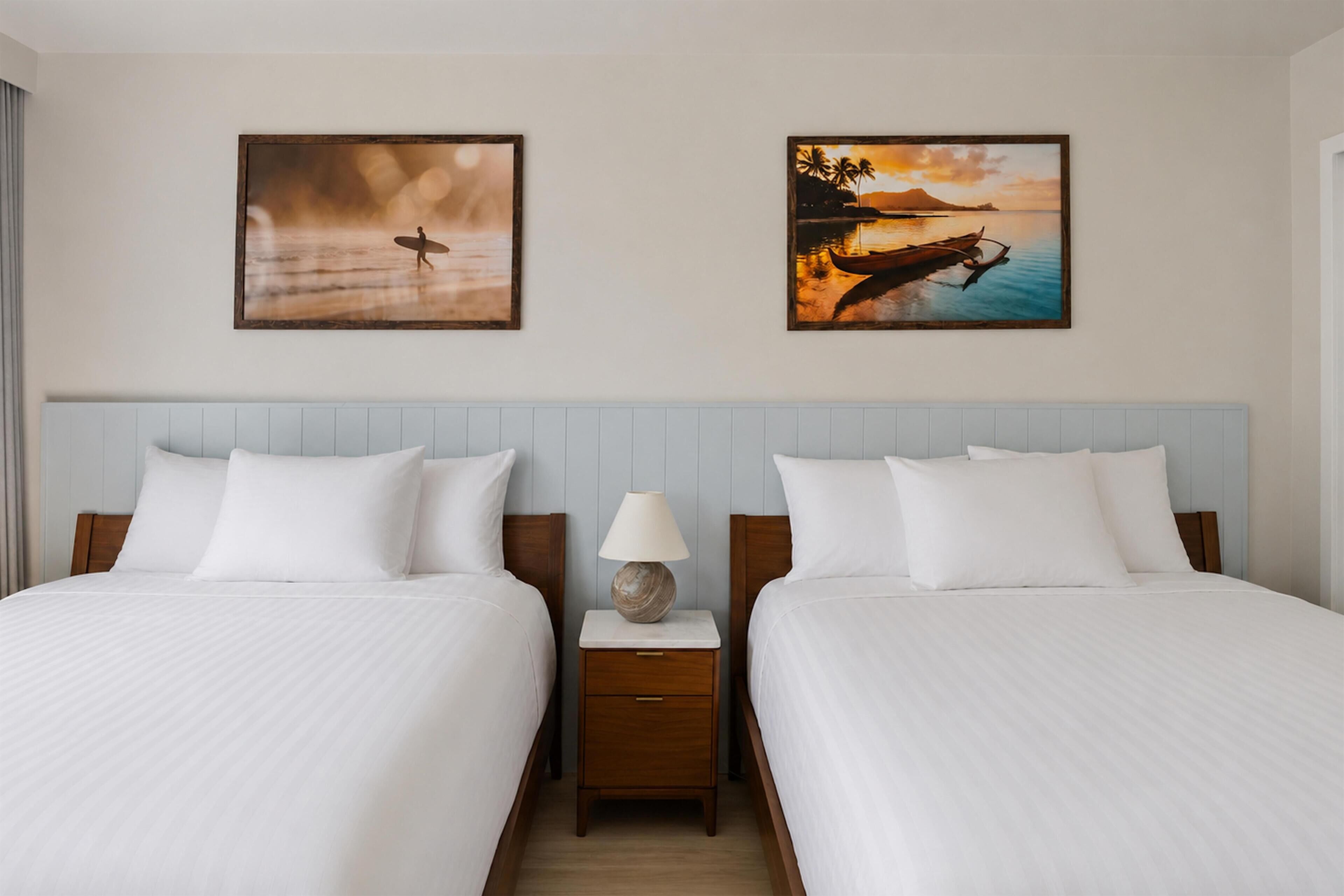 A hotel room with two beds, a nightstand with a lamp, and two framed pictures on the wall.
