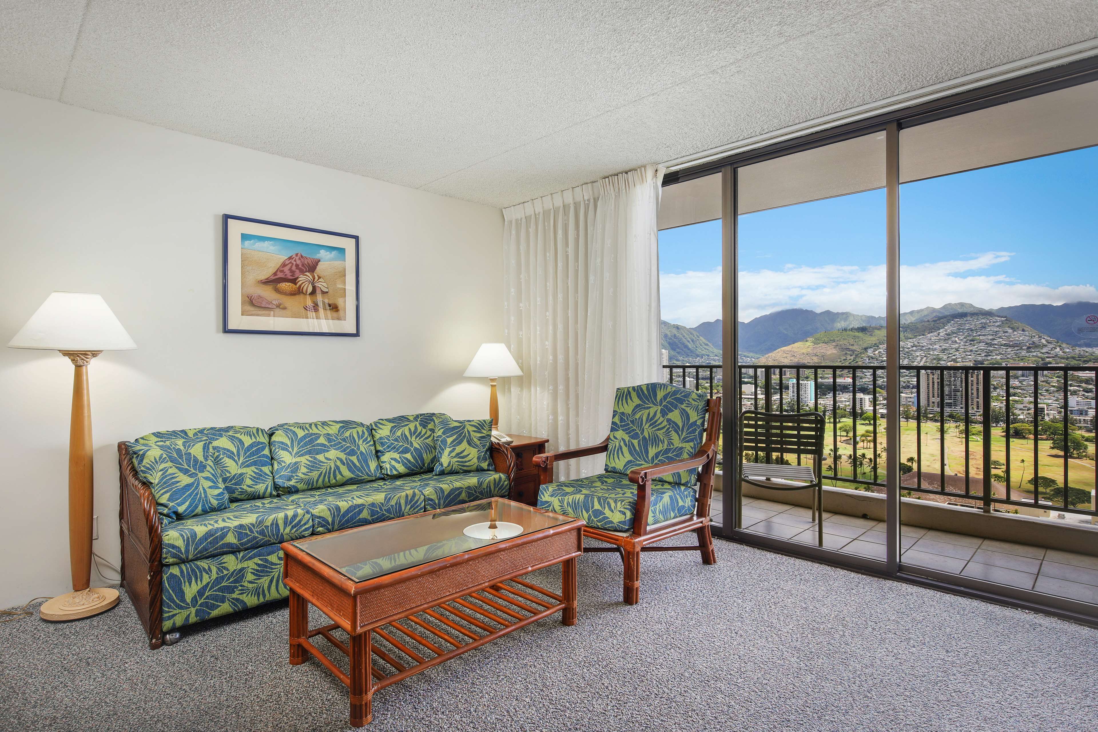 A room with a sofa, armchair, and coffee table, a painting on the wall, and a balcony with a view of mountains and a city.