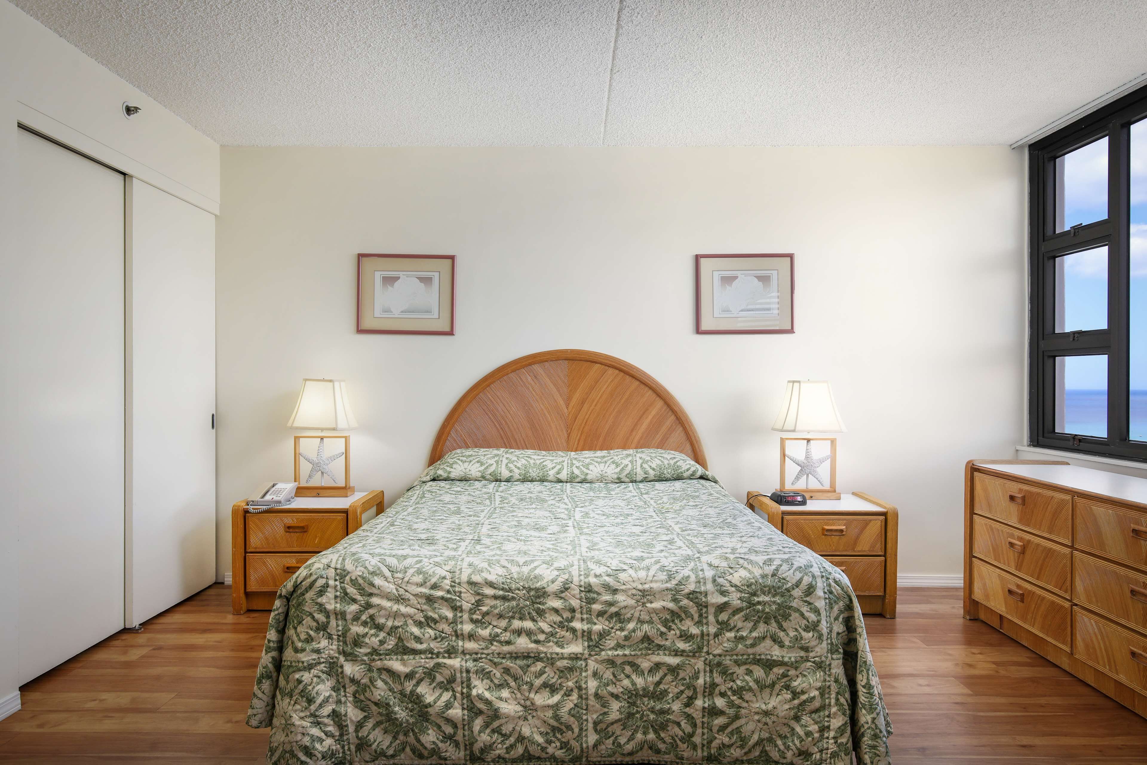 Un dormitorio con cabecera de madera, una cama con colcha floral, dos mesitas de noche con lámparas, un aparador y dos cuadros en la pared.