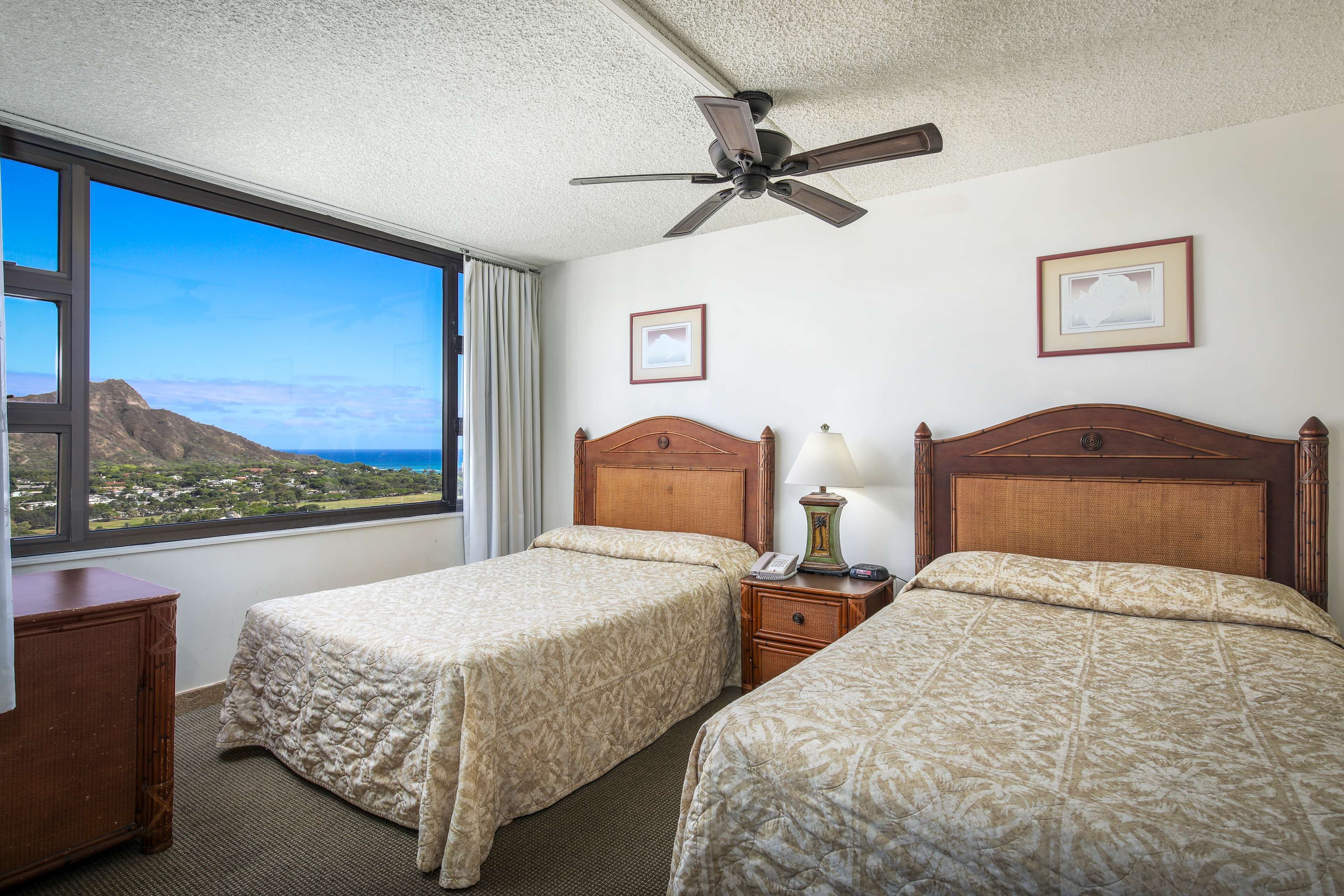 Quarto de hotel com duas camas, um ventilador de teto, mesa de cabeceira com abajur e quadros nas paredes.