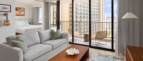 A modern hotel room with a grey sofa, a wooden coffee table, and a balcony with a view of high-rise buildings.