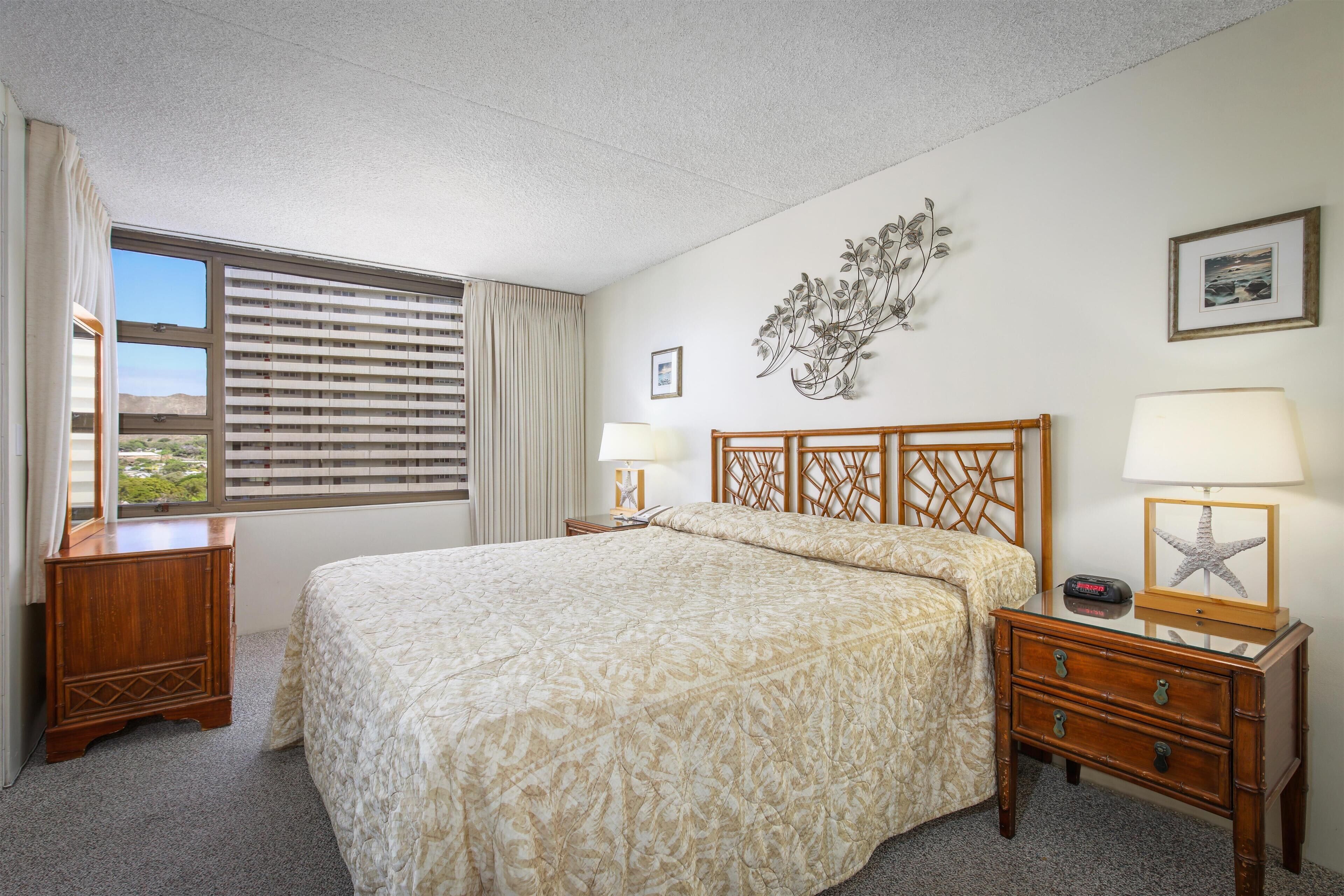 A hotel room with a bed, a nightstand with a lamp, a dresser, a window with blinds, and wall decorations.