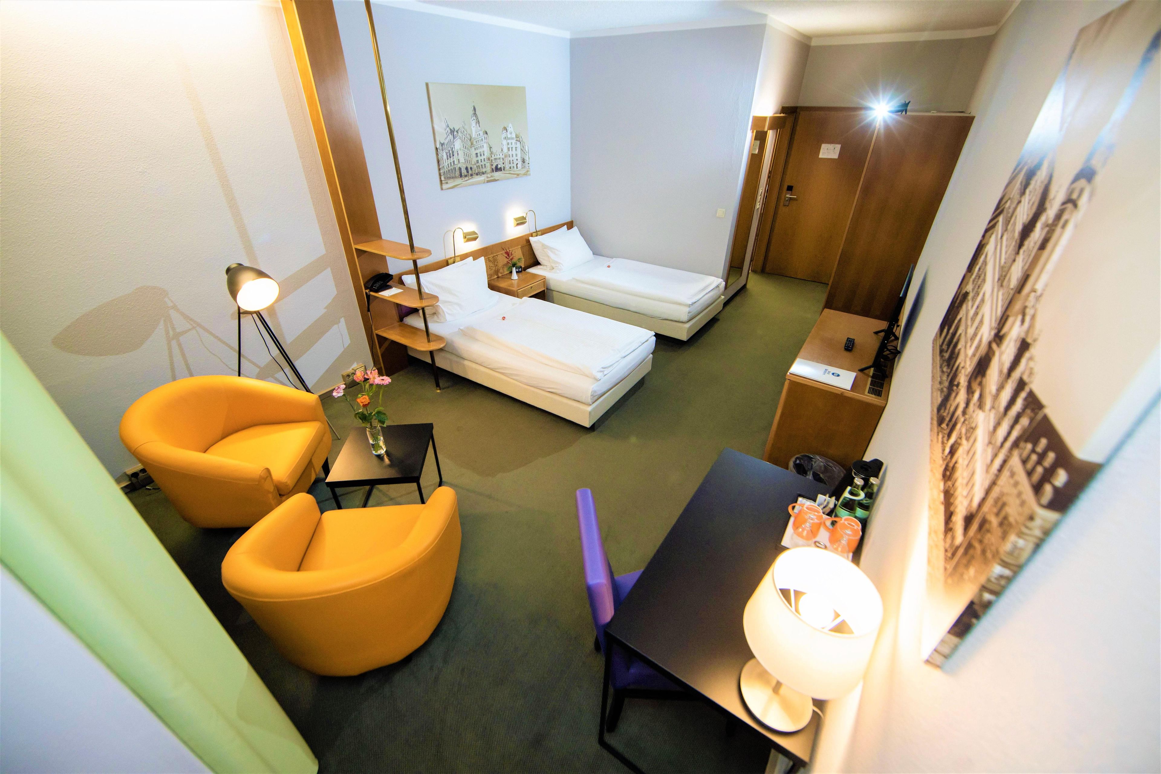 A hotel room with two single beds, a desk, a chair, a small table with a vase, and a lamp.