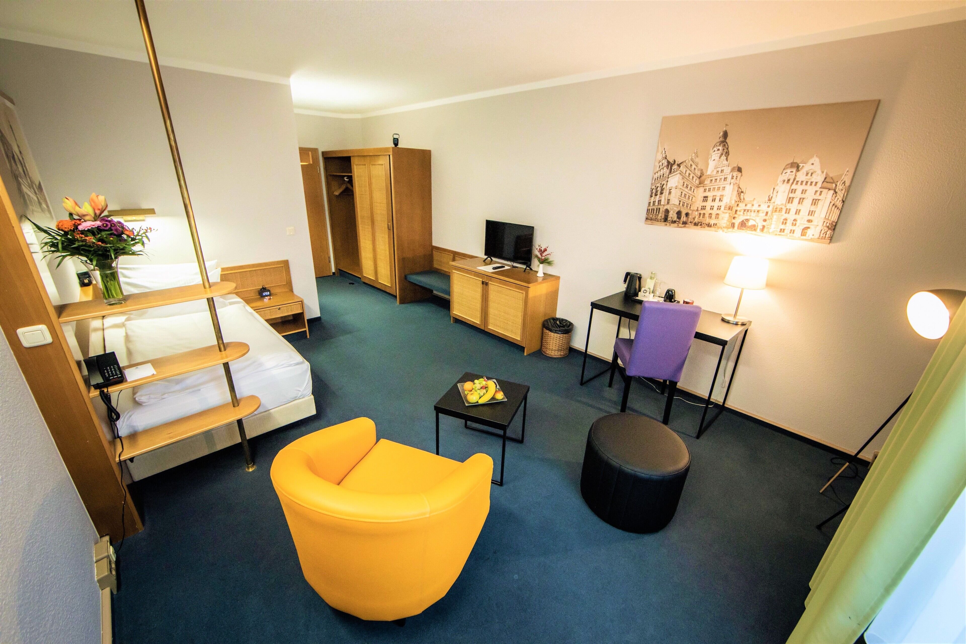 Een hotelkamer met een bed, een gele fauteuil, een klein tafeltje met een lamp, een televisie en een schilderij aan de muur.