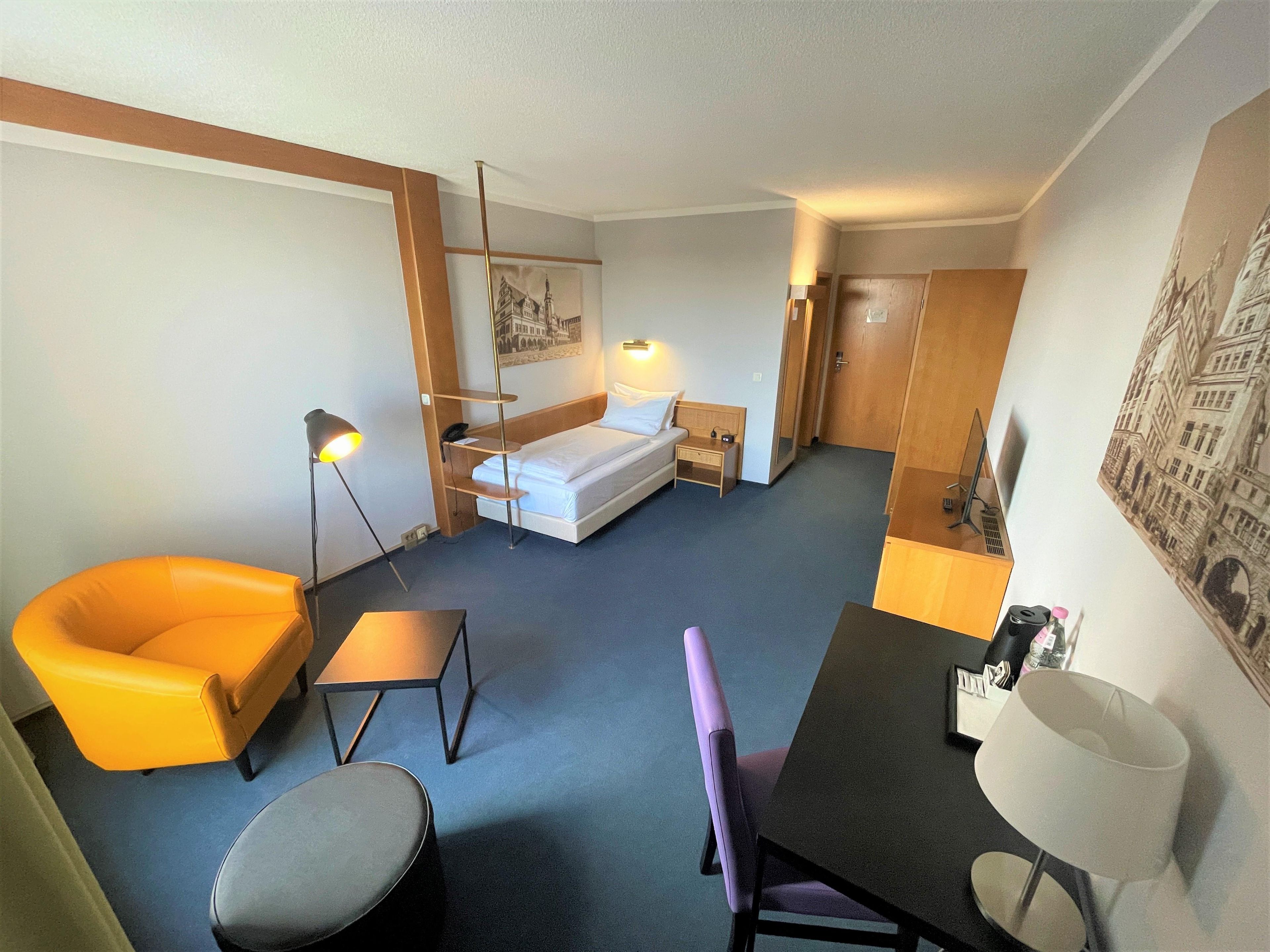 Habitación de hotel con cama, escritorio, silla, mesita, lámpara y un cuadro en la pared.