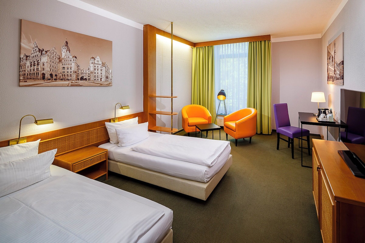 Habitación de hotel con dos camas, un escritorio, una silla, una lámpara y un cuadro en la pared.