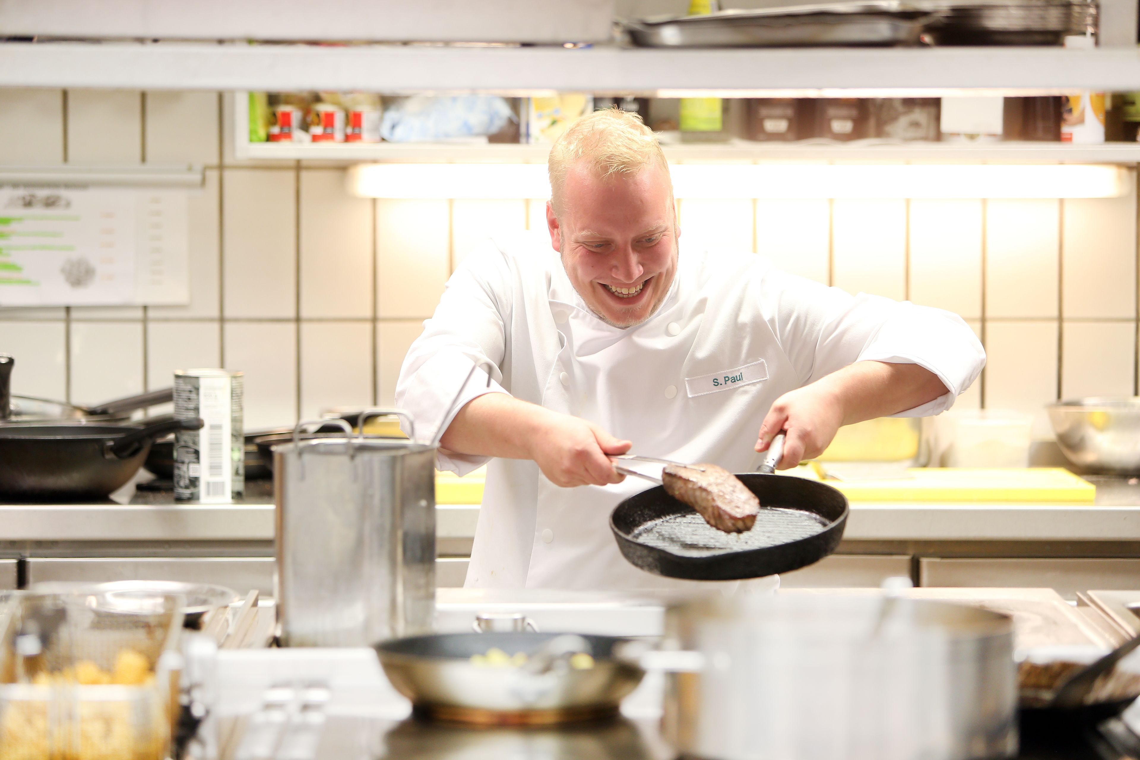 Un chef con uniforme blanco cocinando en una cocina profesional con electrodomésticos de acero inoxidable y un salpicadero de azulejos blancos.