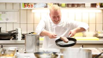 Un chef con uniforme blanco cocinando en una cocina profesional con electrodomésticos de acero inoxidable y un salpicadero de azulejos blancos.