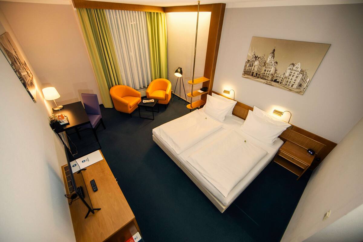 Habitación de hotel con una cama grande, dos sillones naranjas, un escritorio con lámpara y un cuadro de paisaje urbano en la pared.