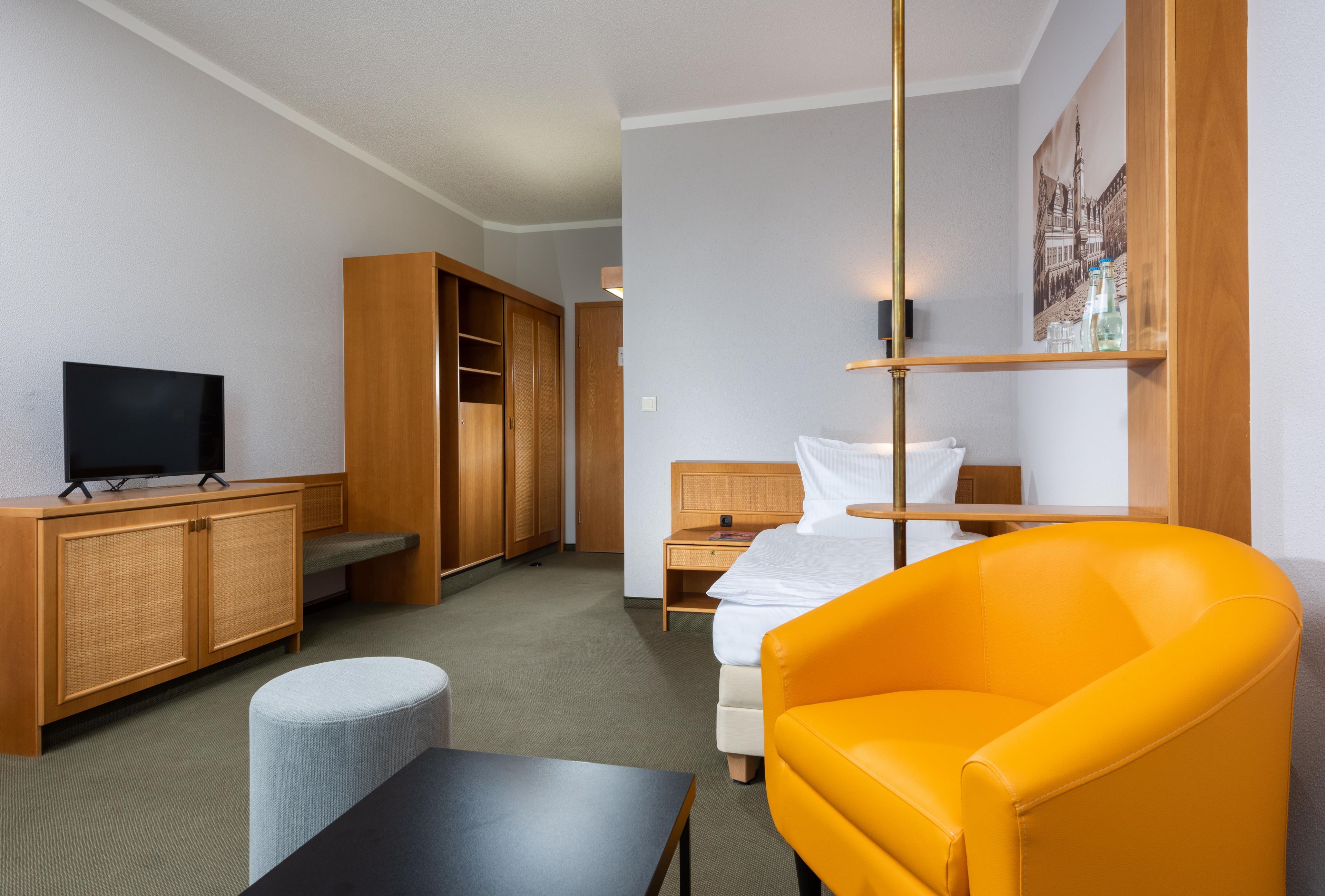 A hotel room with a bed, a desk, a television, a wardrobe, and a bright orange chair.