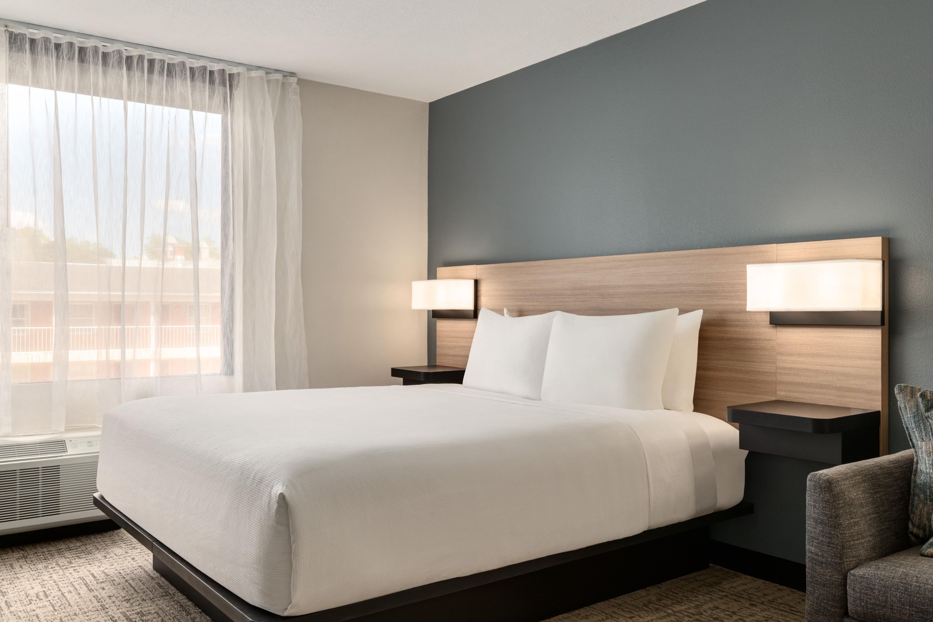 A hotel room with a large bed, two bedside tables with lamps, a chair, and a view of the outside through sheer curtains.