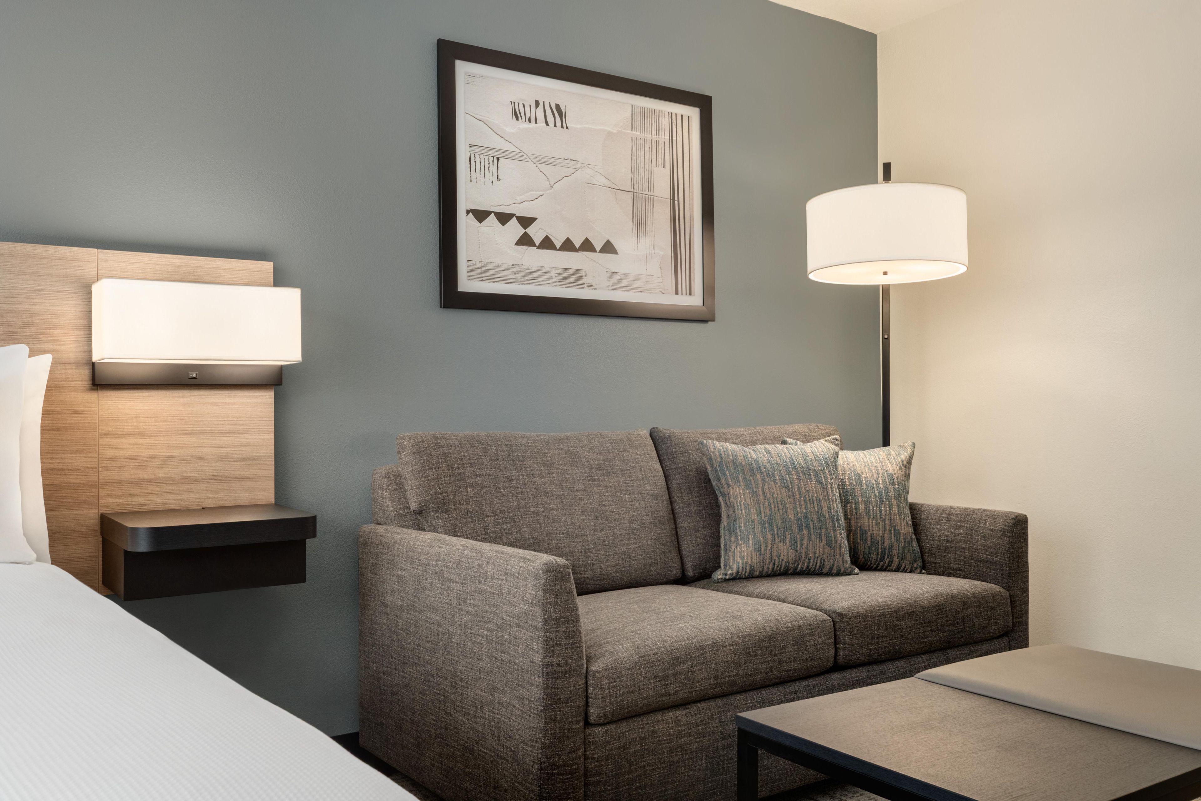 A modern hotel room with a grey sofa, a bedside table with a lamp, a framed abstract artwork, and a bed with white linens.