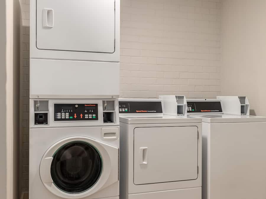 Een wasruimte met gestapelde wasmachines en drogers, bedieningspanelen en een bakstenen muur.