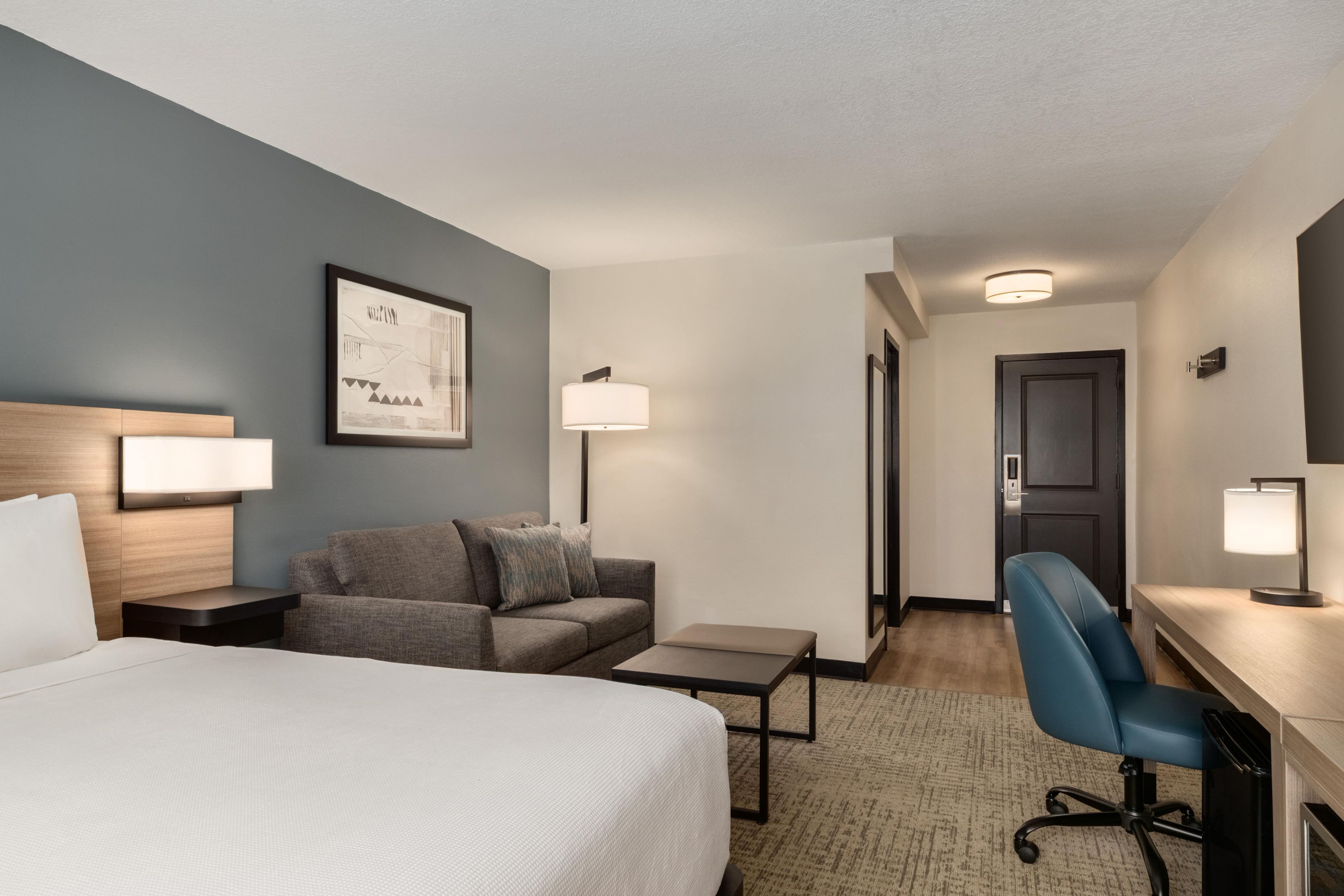 Quarto de hotel com cama, escrivaninha, cadeira, sofá e quadro na parede.