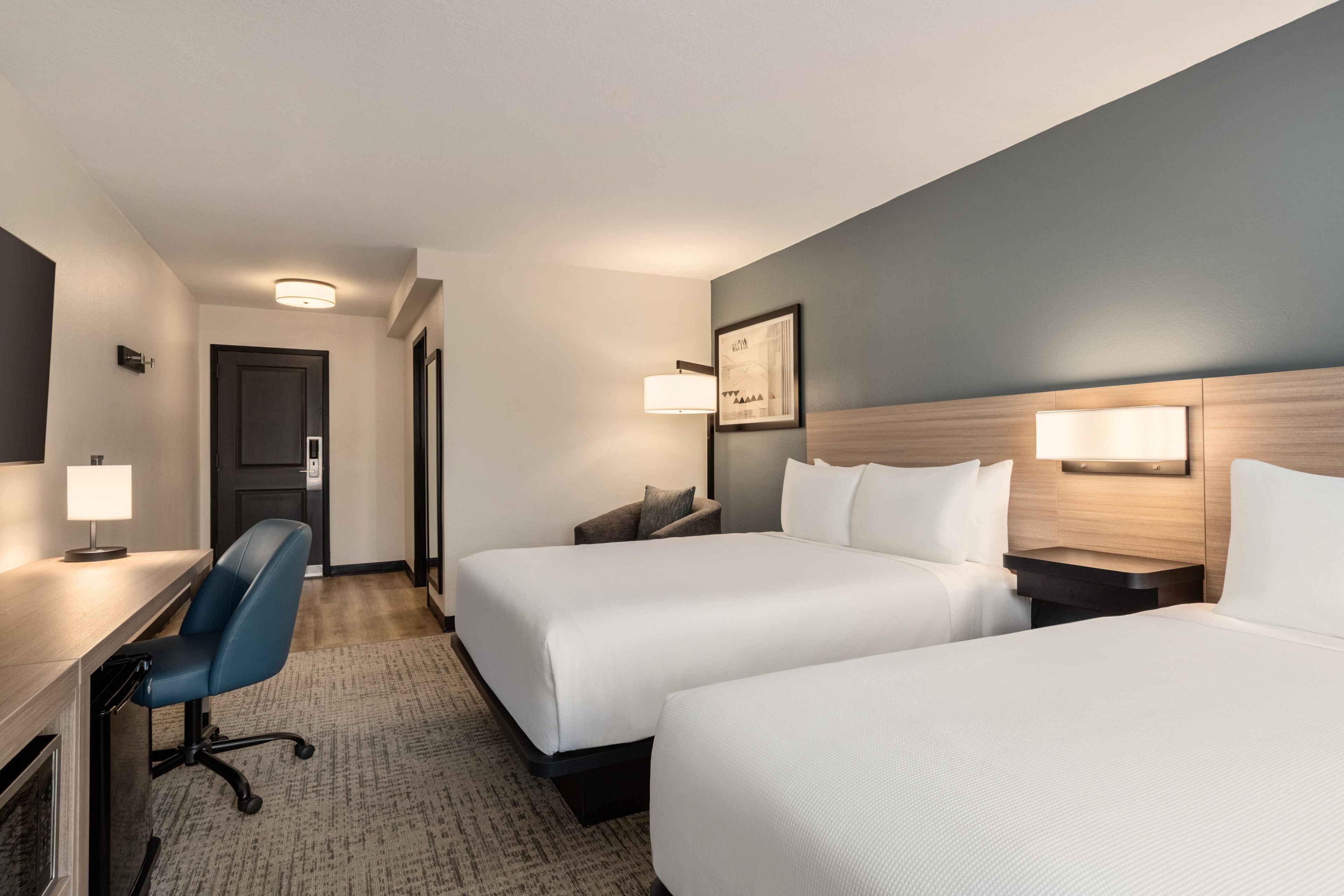 A hotel room with two beds, a desk, a chair, and a door.