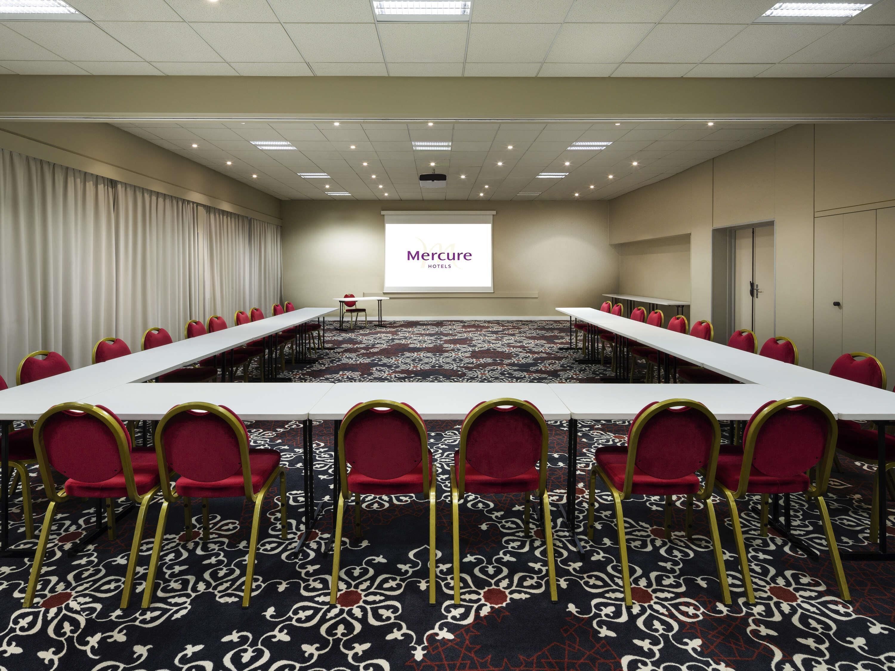 Un salón de conferencias con una larga mesa, sillas rojas, una pantalla de proyector mostrando 'Mercure Hotel' y una alfombra estampada.