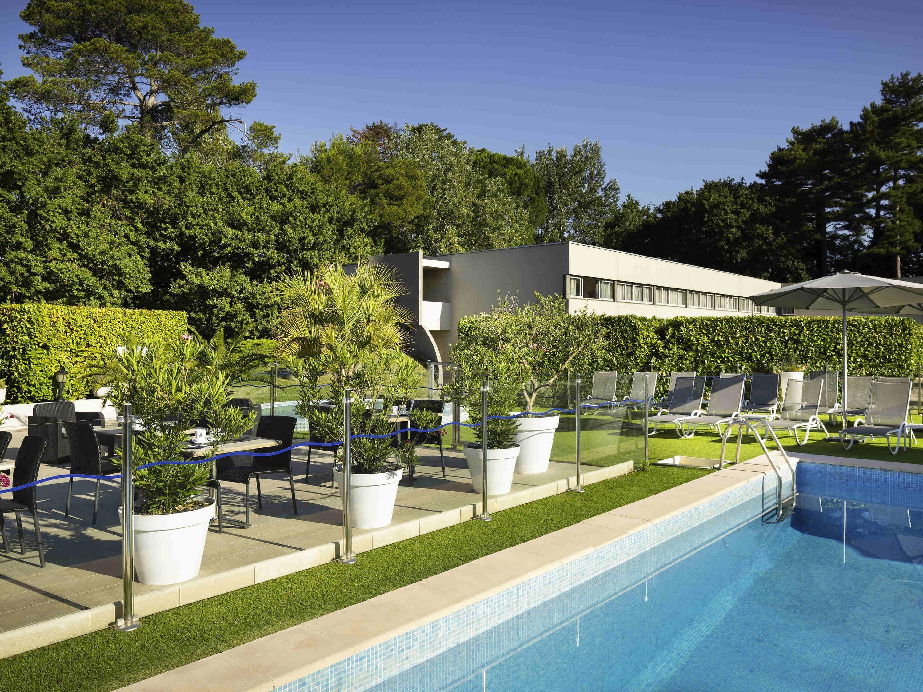 A swimming pool with lounge chairs and potted plants, surrounded by a hedge and a modern building.