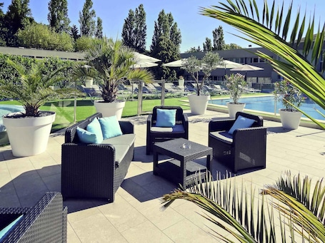 Outdoor seating with potted plants, a pool, and a building in the background.. Mercure Orange Centre