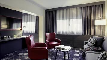 Quarto de hotel moderno com carpete azul escuro em formato hexagonal, um sofá, duas poltronas vermelhas, uma pequena mesa com um livro, uma TV fixada na parede e uma janela grande com cortinas.