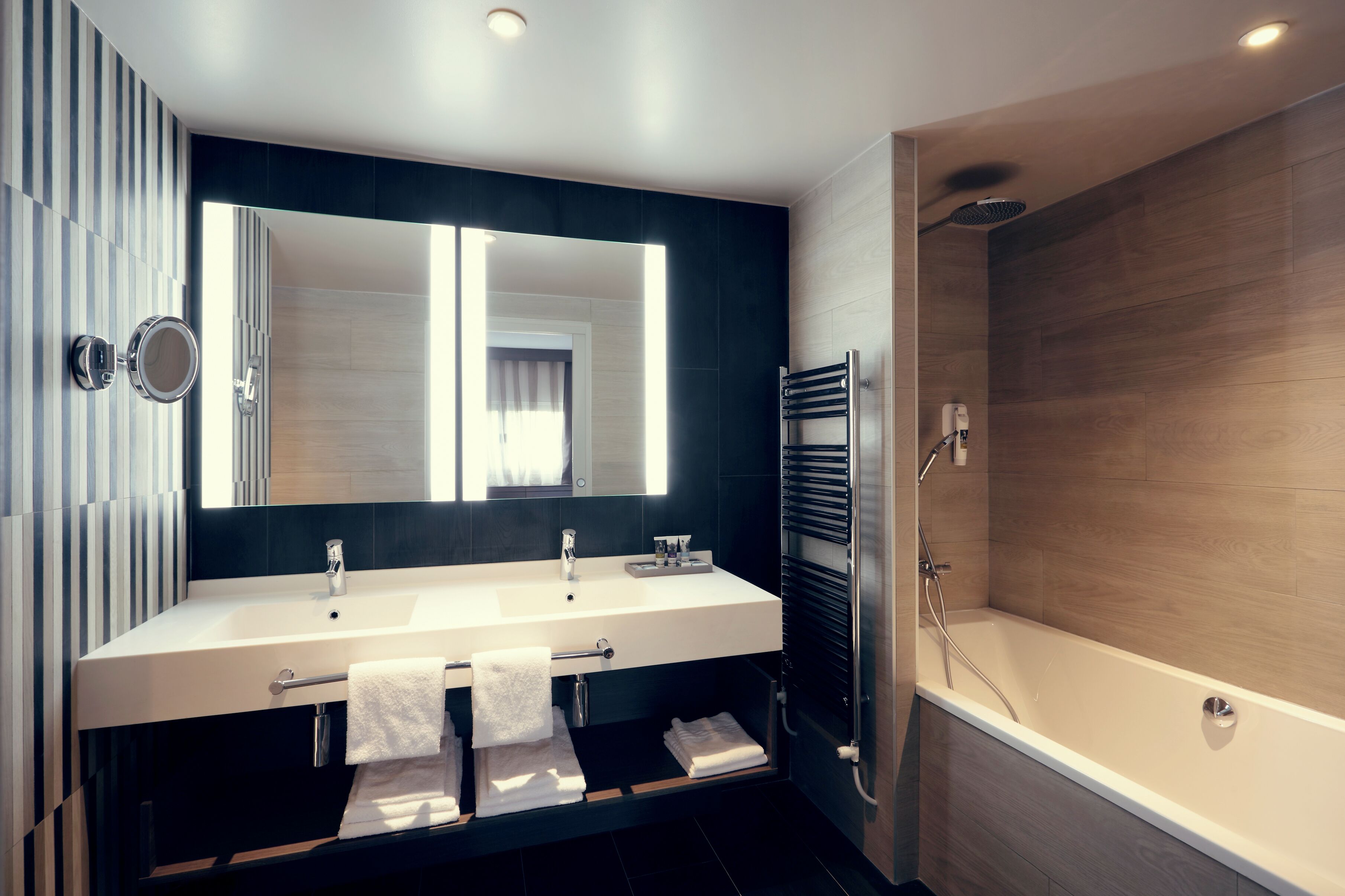 A modern bathroom with a large mirror, two sinks, a towel rack, and a bathtub.