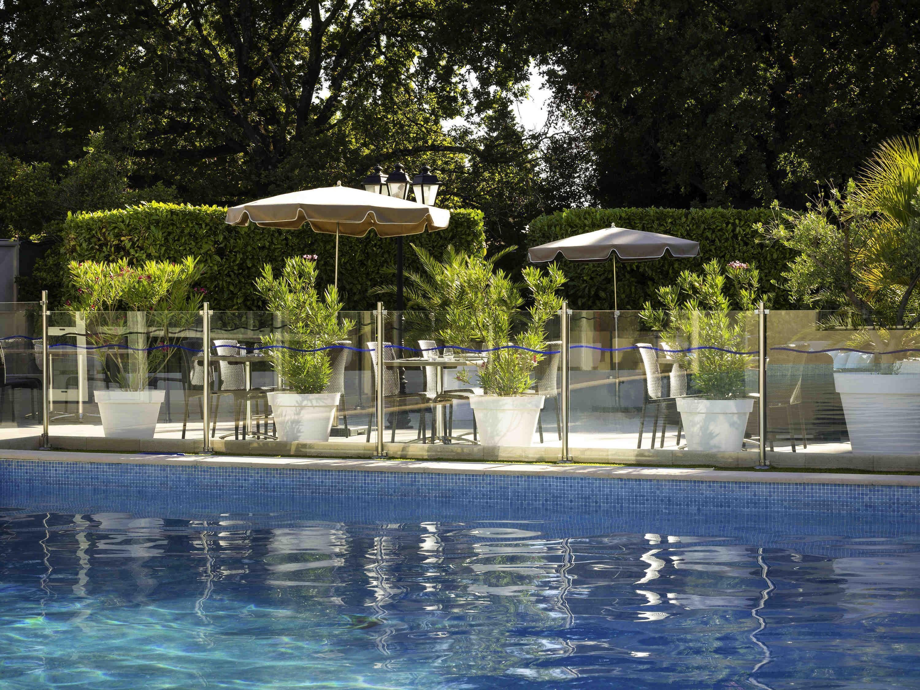A swimming pool with a tiled edge, surrounded by outdoor seating with umbrellas, potted plants, and a glass fence.