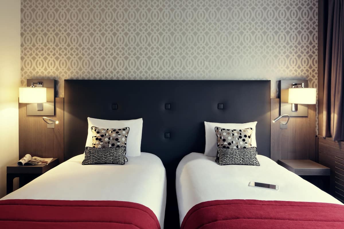 Habitación de hotel con dos camas, cabecera oscura, papel tapiz estampado, lámparas de noche y una manta roja.