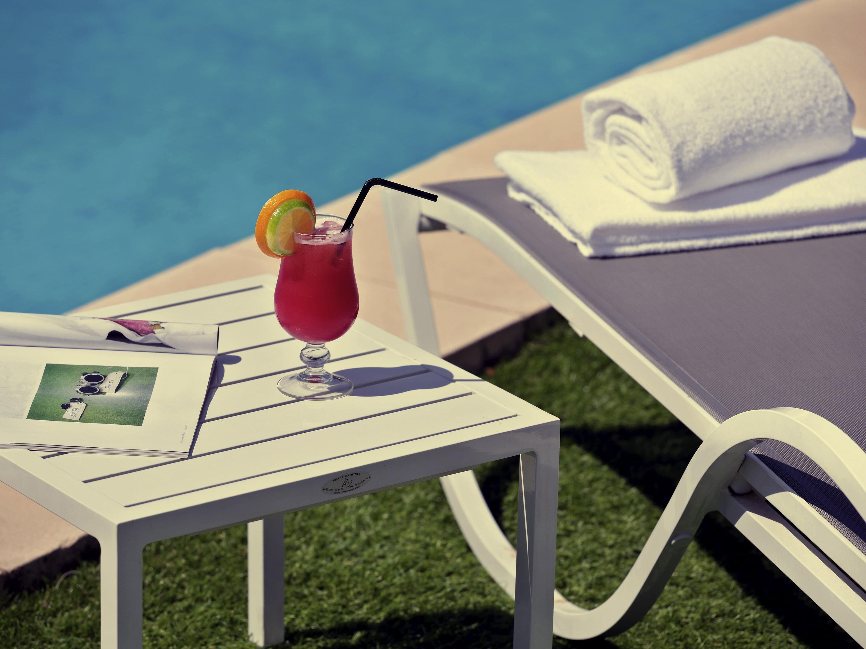 Una silla de salón junto a la piscina, un vaso de bebida roja con pajita y decoración, y una toalla enrollada sobre una mesa blanca.