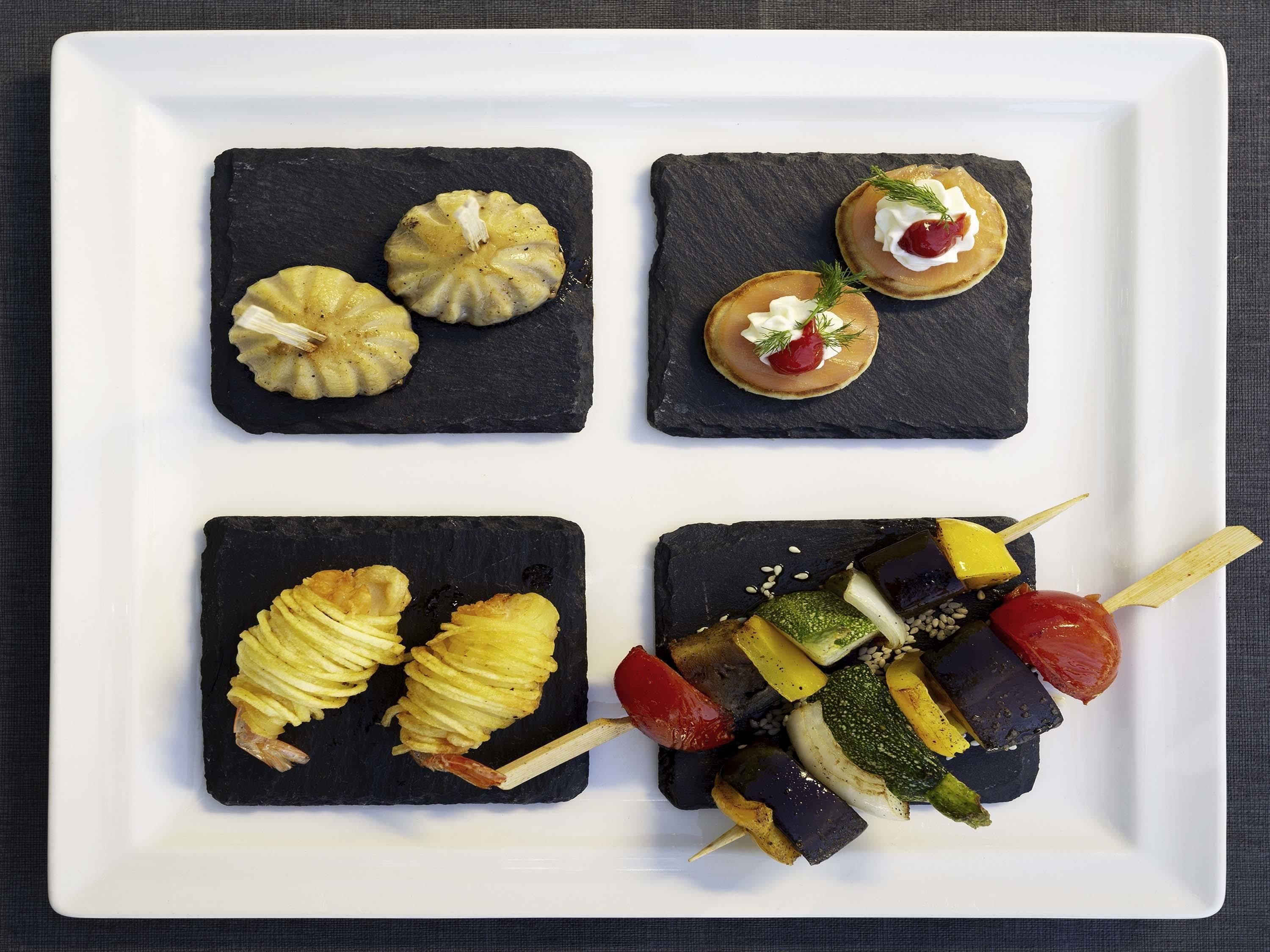 Un plato blanco cuadrado con cuatro baldosas negras de pizarra, cada una con diferentes alimentos.