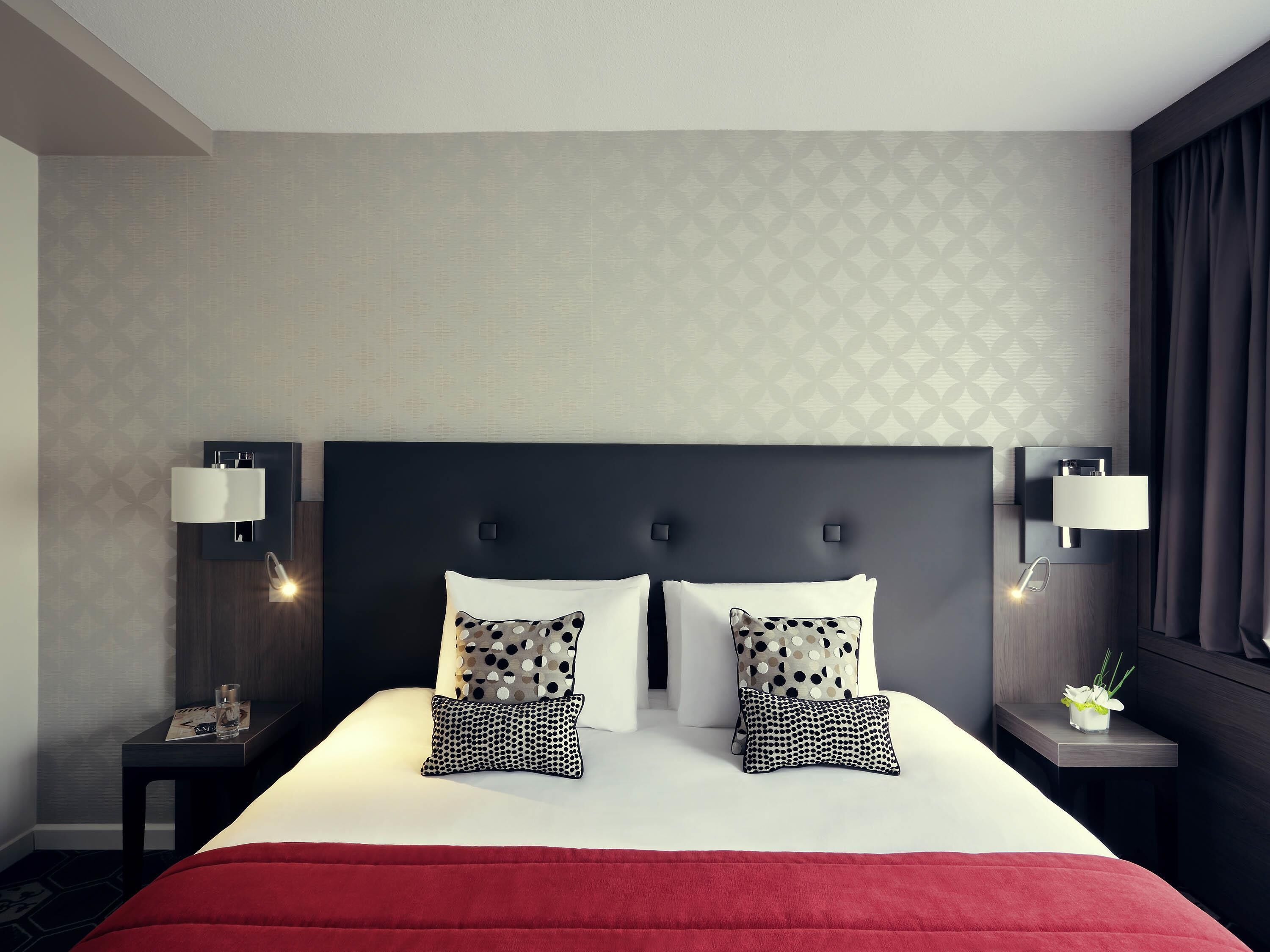 A hotel room with a large bed, two bedside tables, a nightstand, a red bedspread, and patterned wall decor.