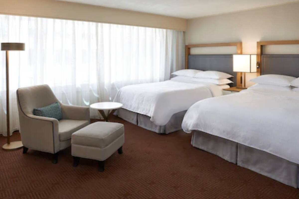 A hotel room with two beds, a chair, a small table, and a lamp.