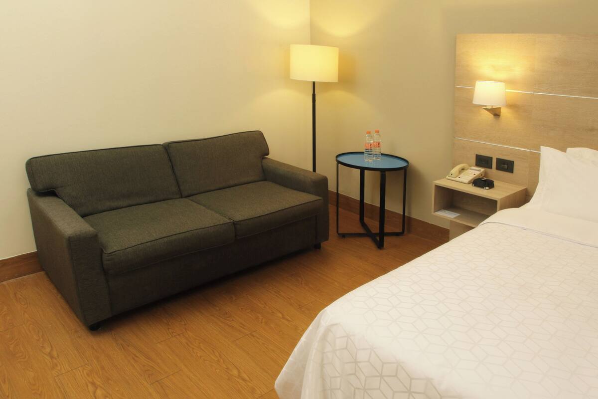 A hotel room with a bed, a sofa, a small table with a lamp, and a nightstand with a phone and a bottle.
