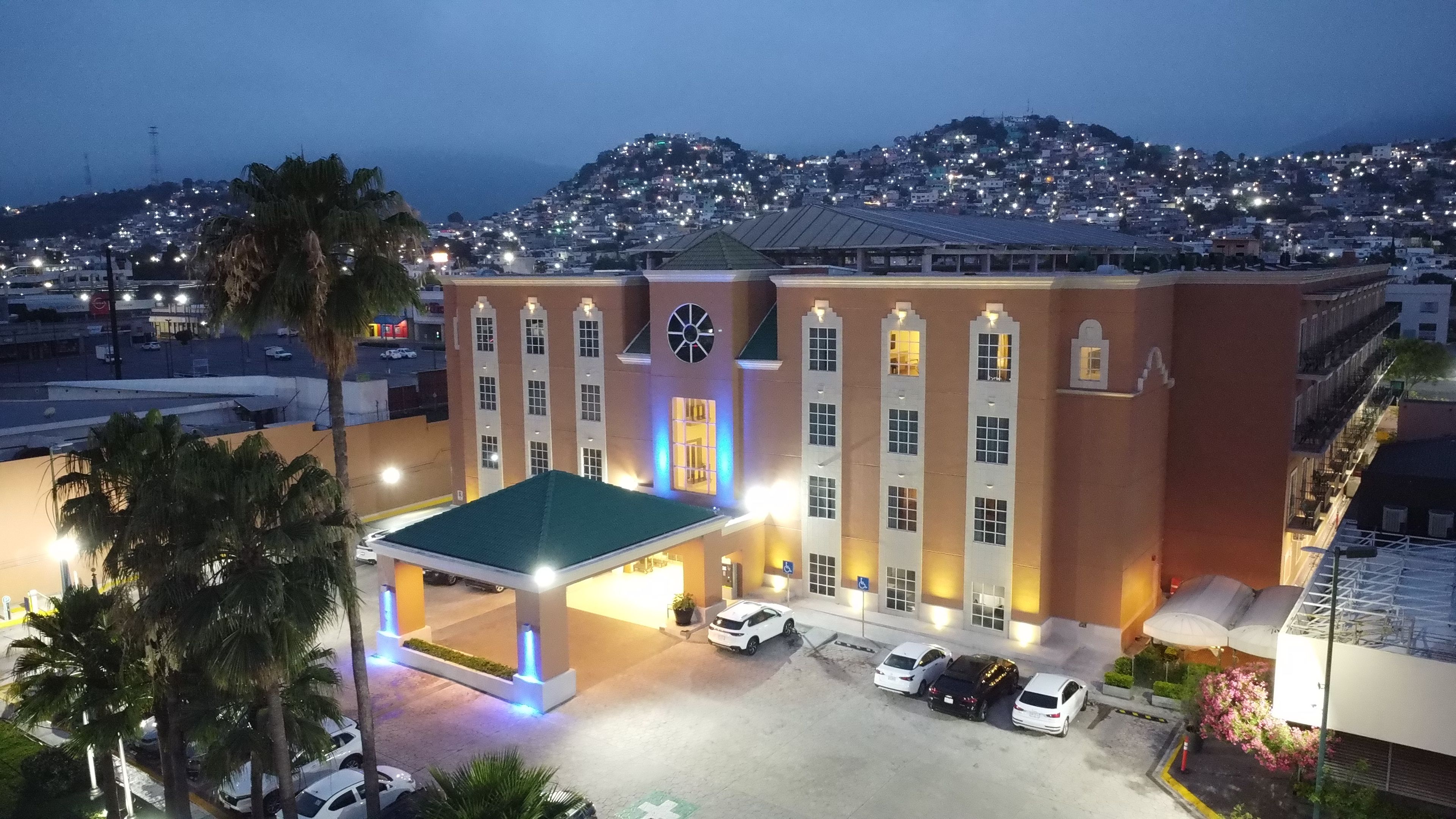 Un hotel amb un edifici central, una zona de pàrquing coberta i vista a una vila situada en un turó.
