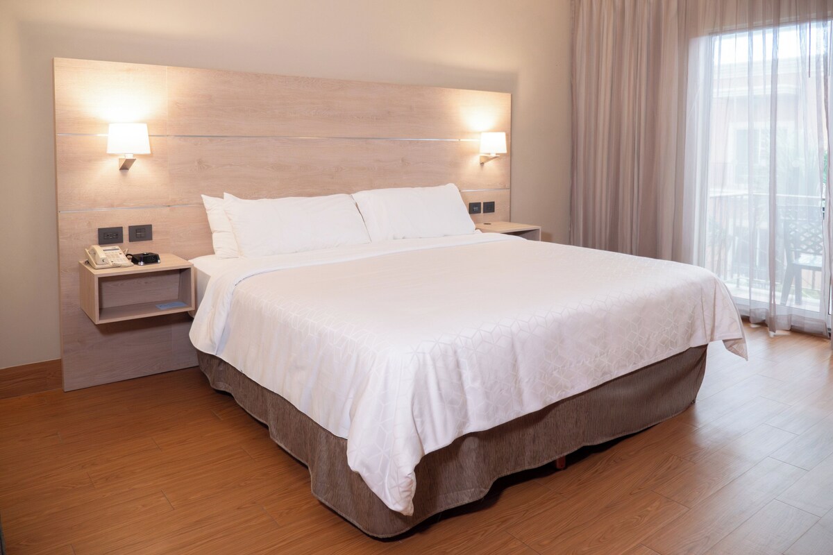 A hotel room with a large bed, bedside lamps, a phone on the nightstand, and a view of the outdoors through sheer curtains.