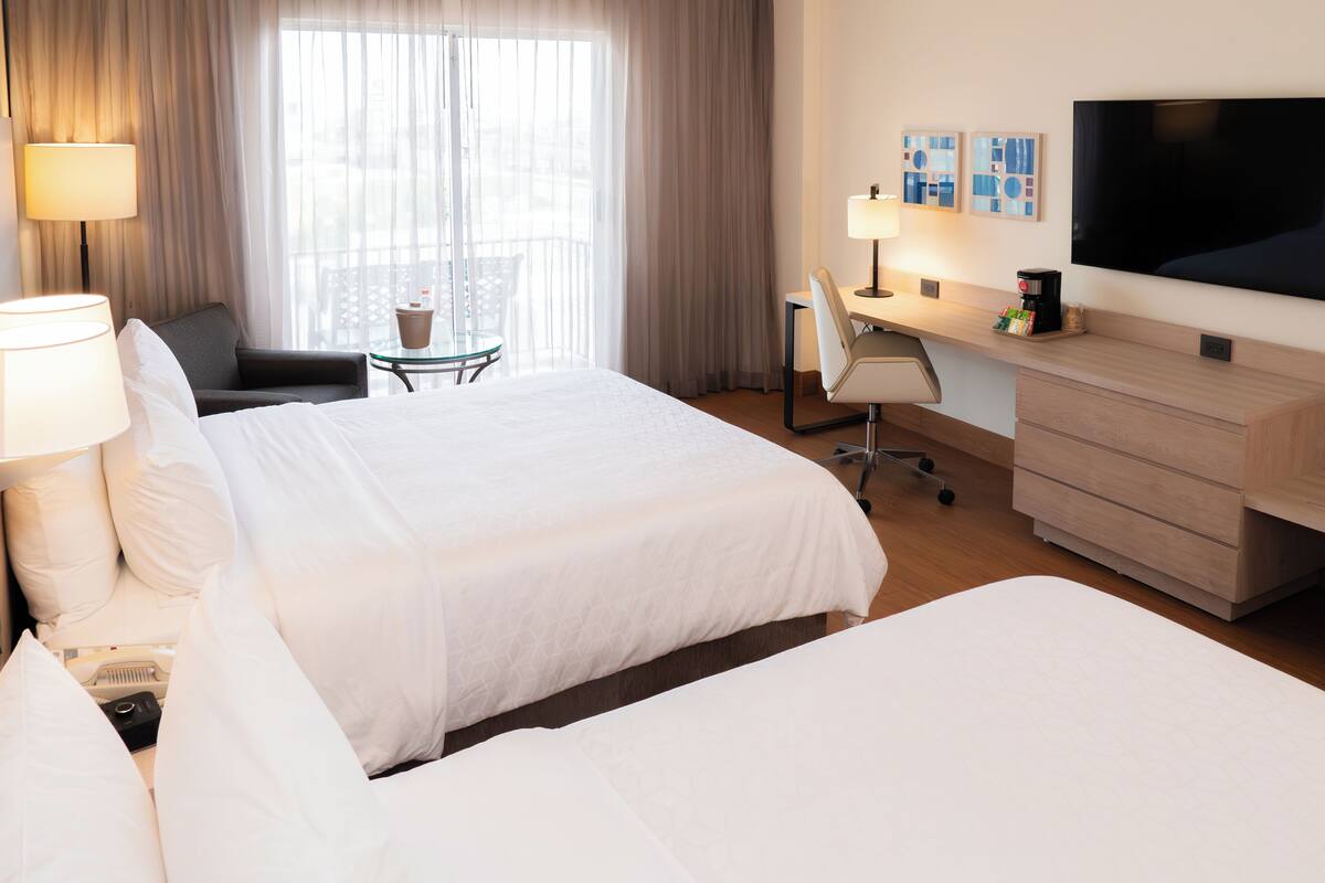 A hotel room with two beds, a desk, a chair, a TV, and a balcony with curtains.